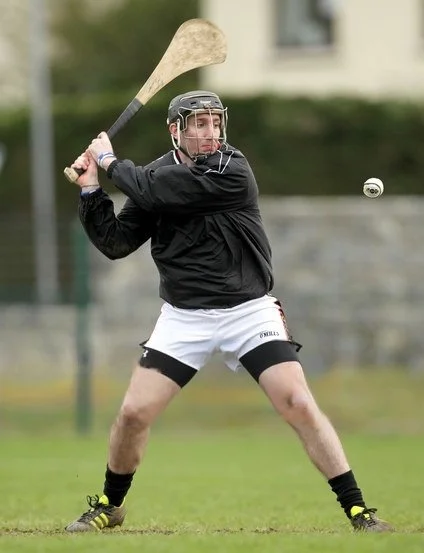 A man in black jacket, white shorts, and a helmet playing hurling, preparing to hit a sliotar with a hurley during a game.