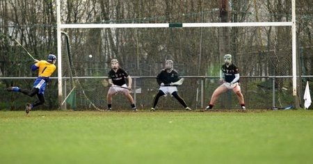 A rugby game with one player attempting to kick the ball while three players in front try to block or defend, on a grassy field with goal posts.