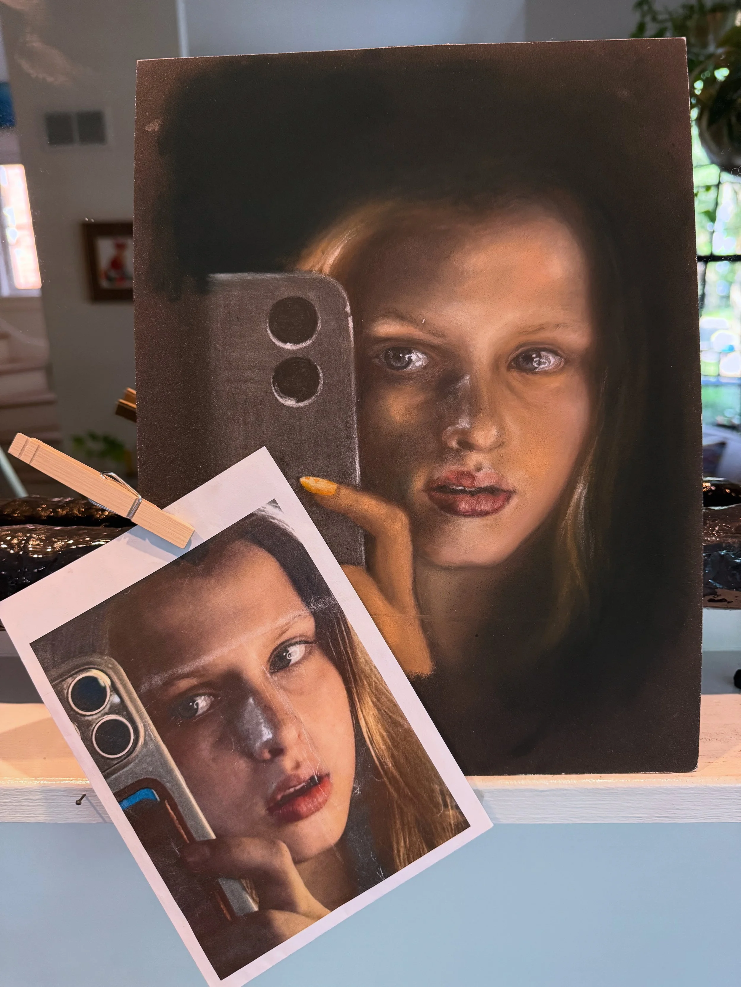 A realistic portrait of a young woman with long hair, holding a smartphone, with a printed reference photo of her face and phone in front of the painting.