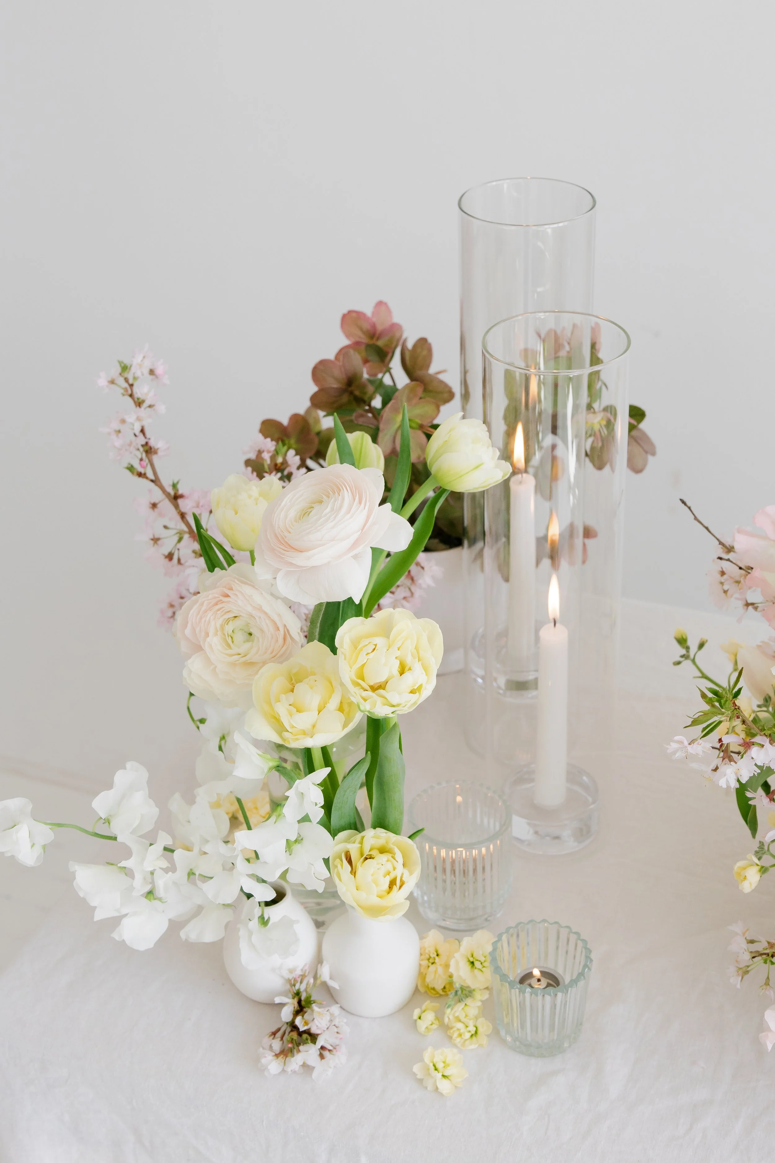 A floral arrangement on a white table includes white ranunculus, pale yellow tulips, pinkish-brown leaves, and white cherry blossoms. The arrangement is complemented by glass candle holders with lit candles, set against a plain white background.