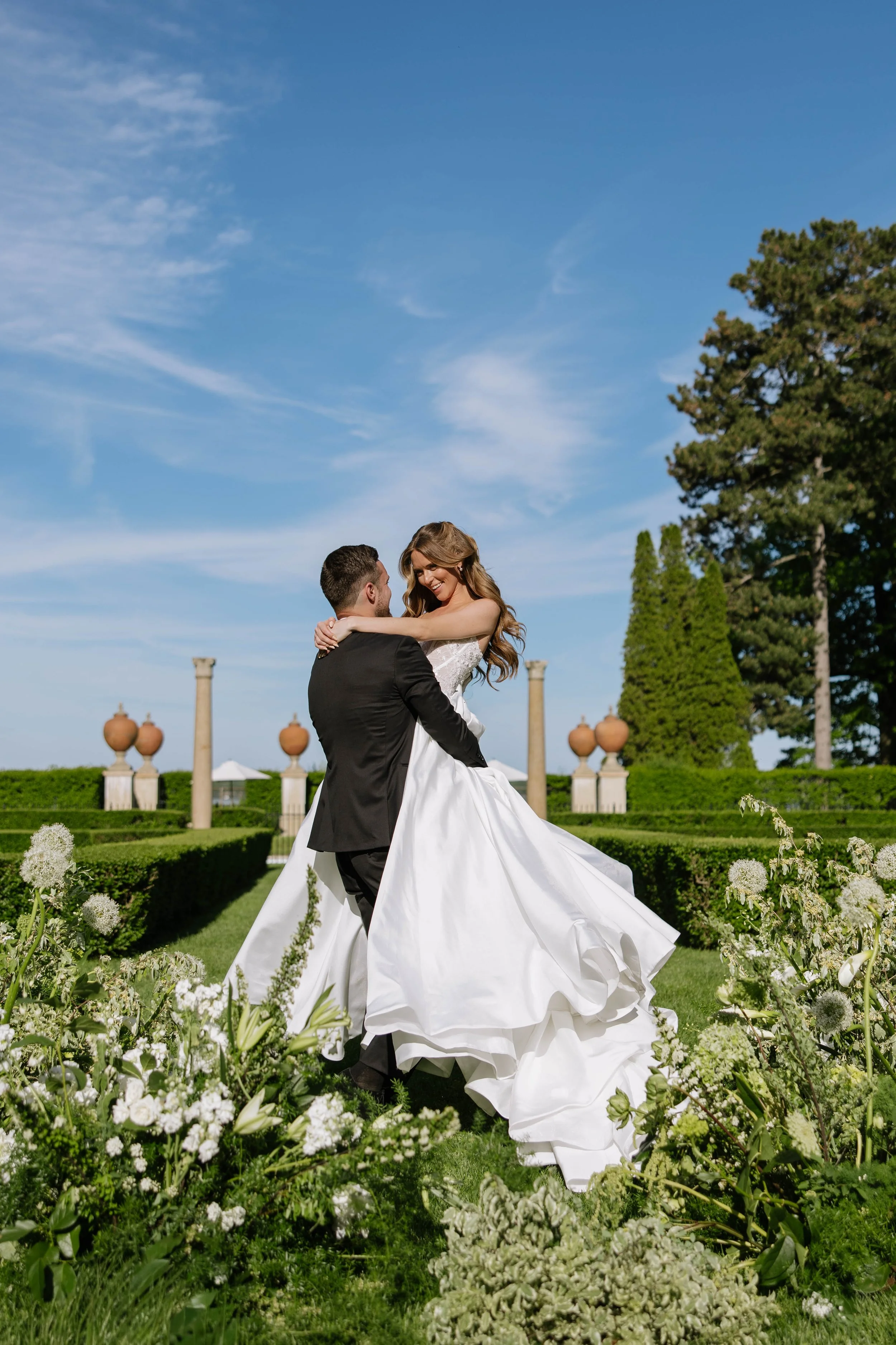 A bride and groom wedding photo in a garden with a blue sky in Geneva. The groom is lifting the bride who is smiling. They are surrounded by white allium, hydrangea, delphinium and greenery, with tall trees and columns in the background.