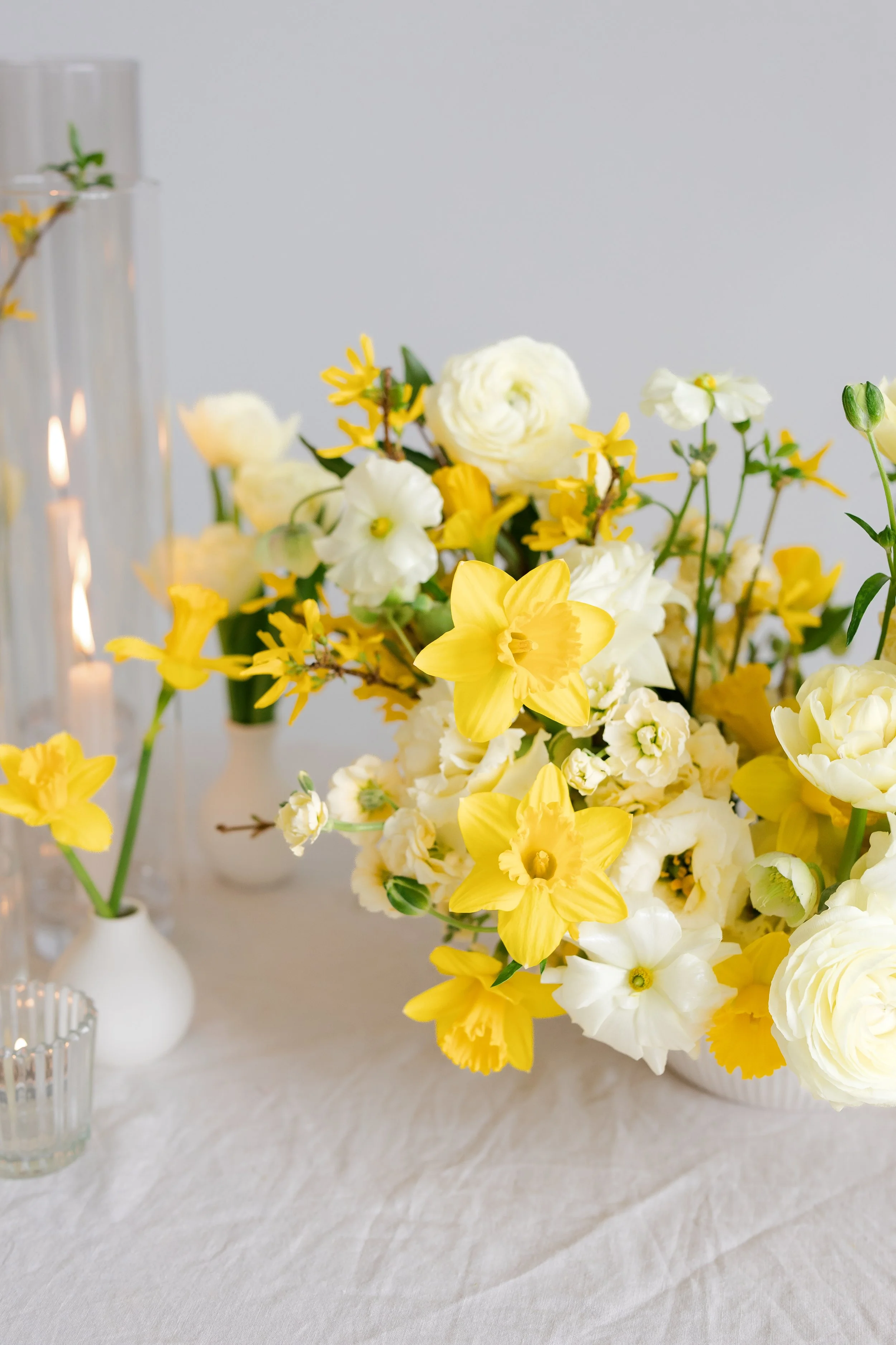 A floral arrangement with white and yellow flowers in a white vase on a table with a white tablecloth. Tall glass candle holders are visible on the left side of the image for a wedding ceremony in buffalo, ny.