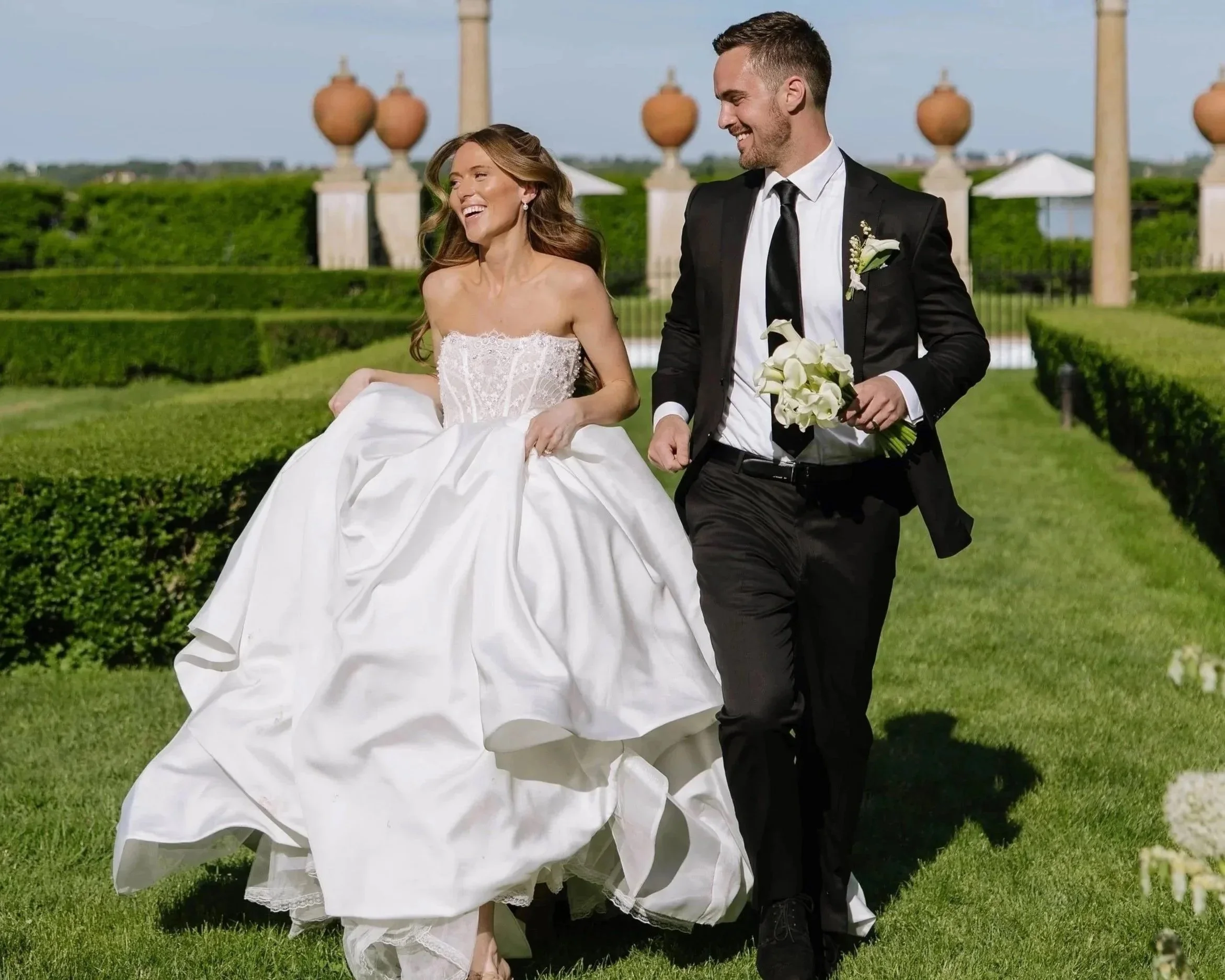 A smiling bride in a white wedding dress holding her gown and a smiling groom in a black suit holding a bouquet of white calla lilies, walking together outdoors on a sunny day with decorative columns and green hedges in the background.