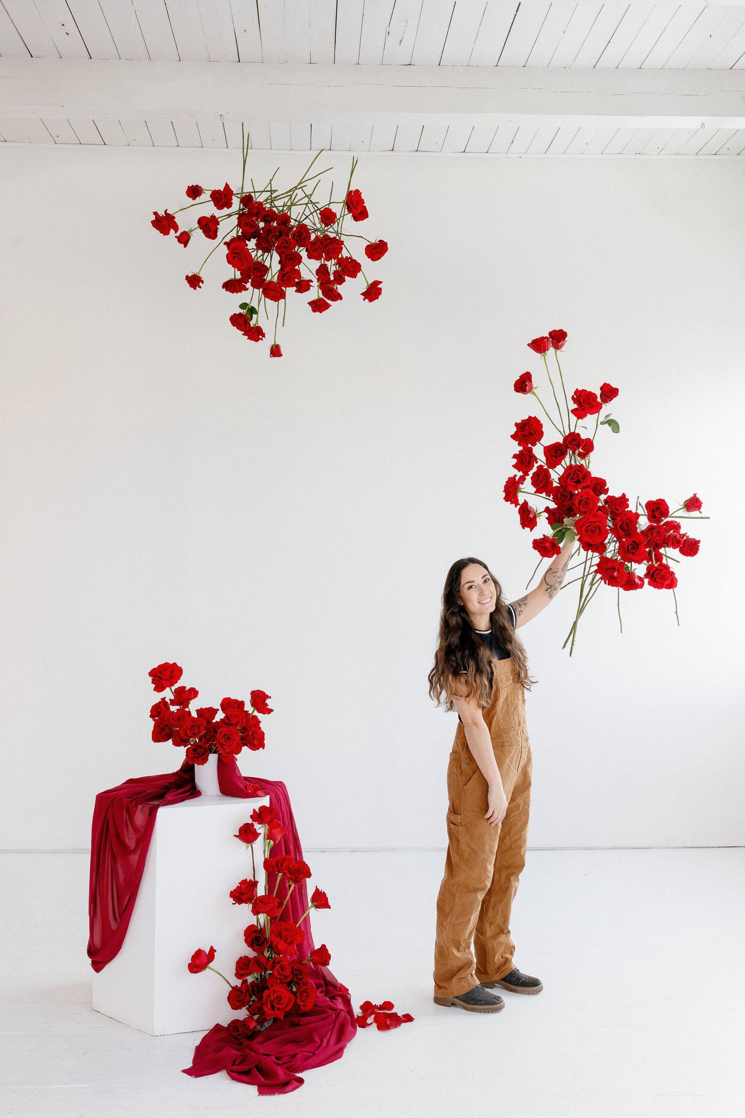 A woman with long, wavy hair and tattoos on her arm stands next to a display of red roses. She is holding a large bouquet of roses with his arm extended upward. The display includes a white pedestal with a bouquet of roses on top, decorated with red 