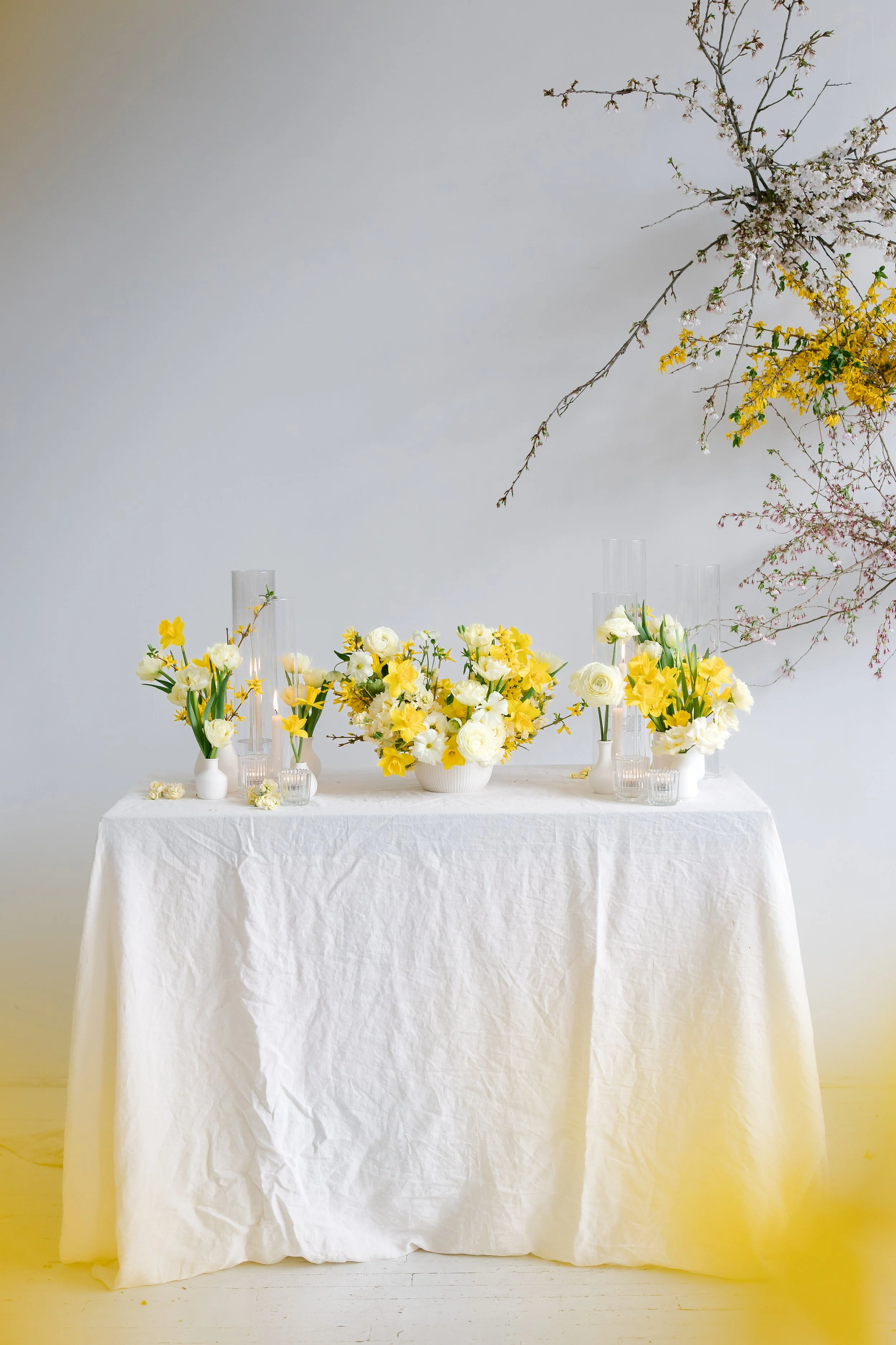 Arrangement of white and yellow flowers on a table covered with a white tablecloth, with tall glass vases and a branch of cherry blossoms in the background.