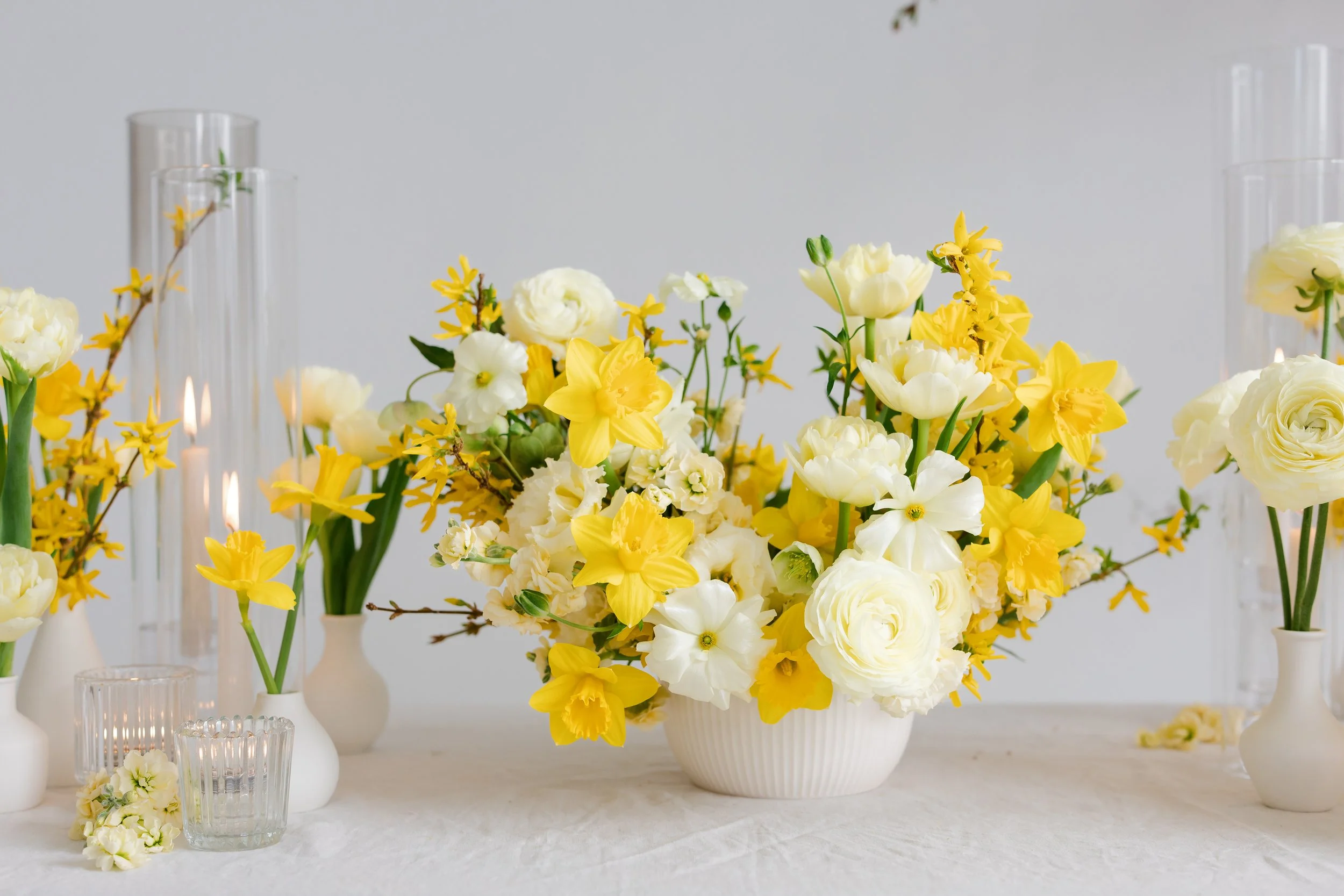 A floral arrangement with yellow and white flowers in a white ceramic vase, surrounded by smaller white vases and glass containers on a white tablecloth against a plain light gray background.