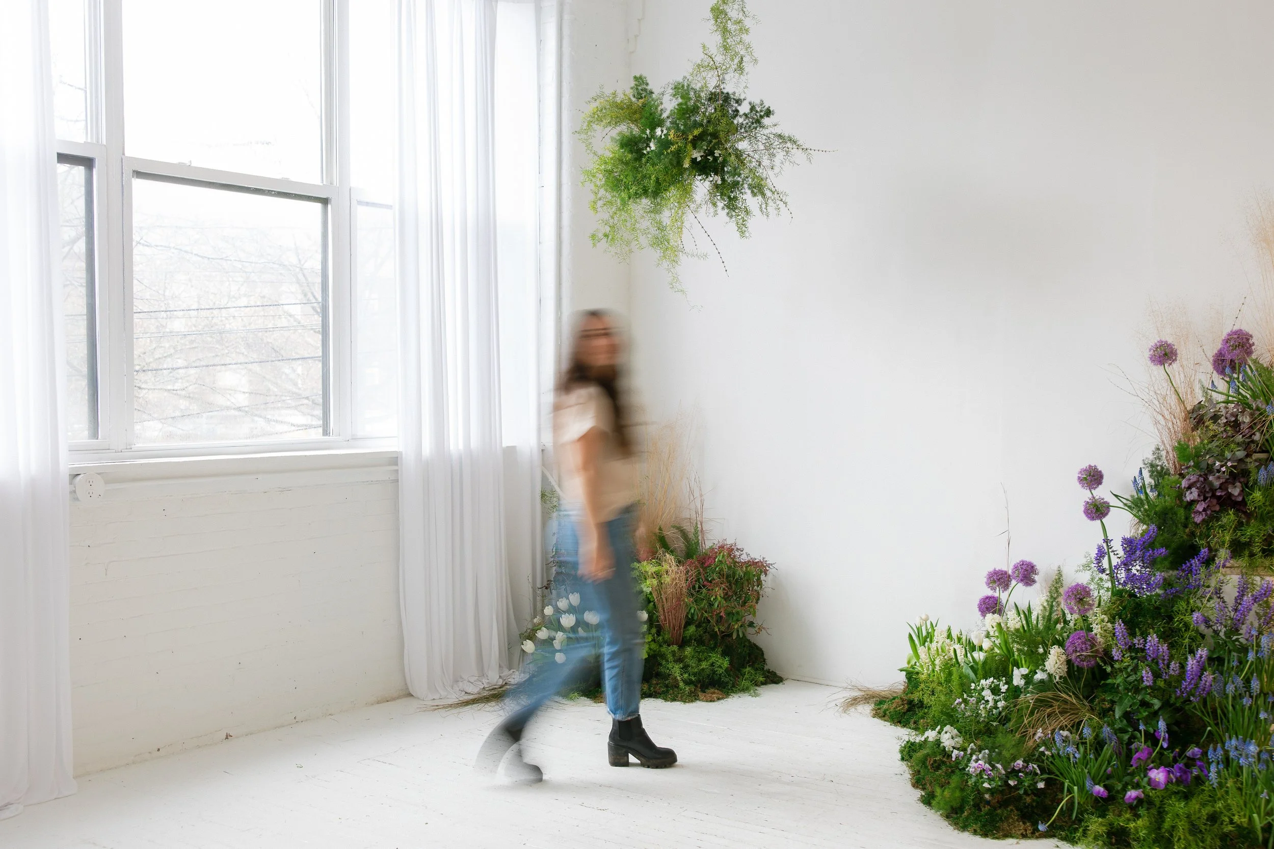 A blurry woman in a white top, blue jeans, and black boots walking past a large indoor display of purple alliums, salvia,  white tulips and hyacinth, and moss with textured greens suspending in the air against a bright white studio with a window.