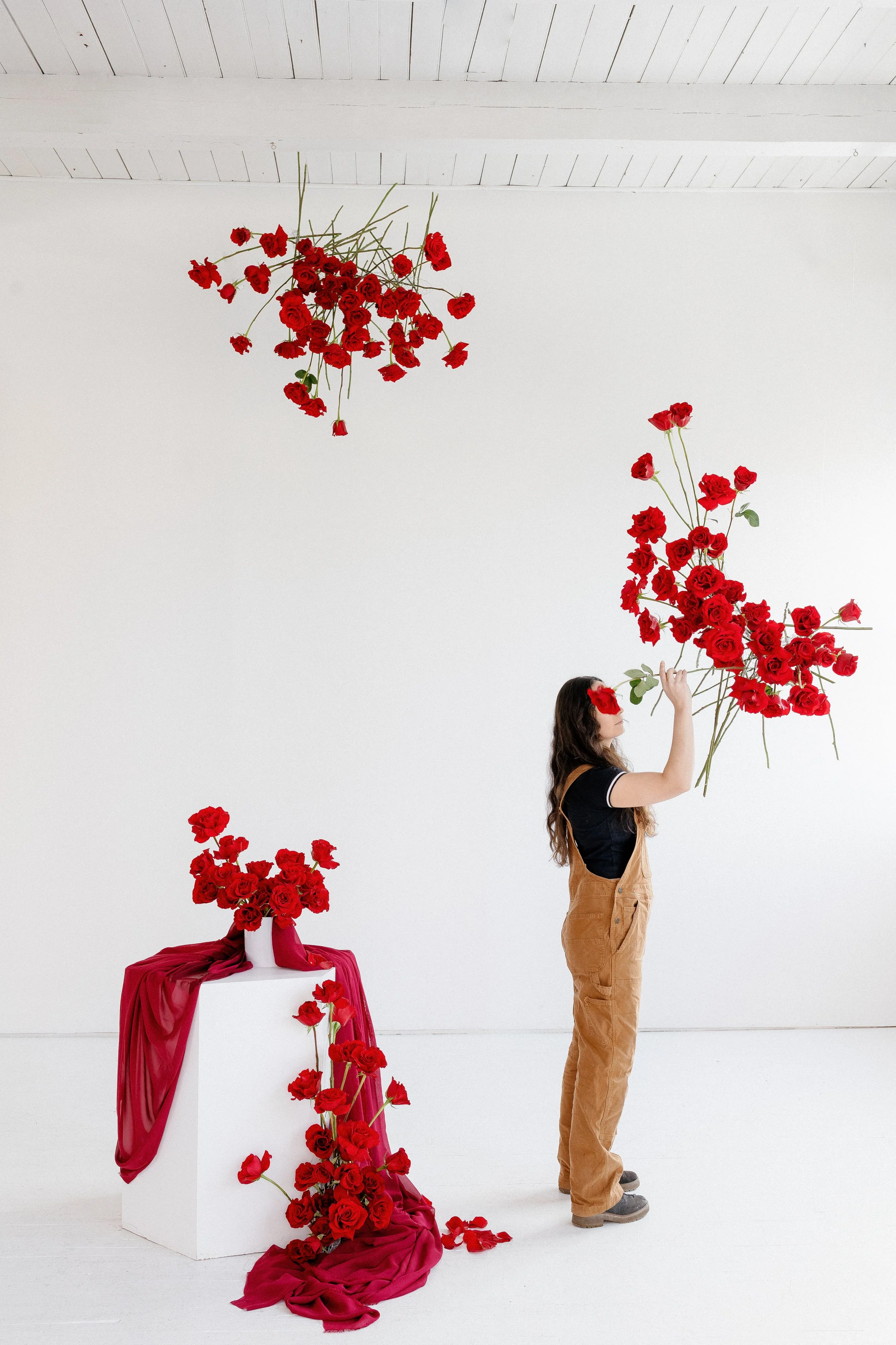 A woman with long dark hair and wearing brown overalls holds a large bunch of red roses in a white art studio with a white wall and wooden ceiling. A table draped with red fabric and decorated with more red roses stands to her left, and roses are sca