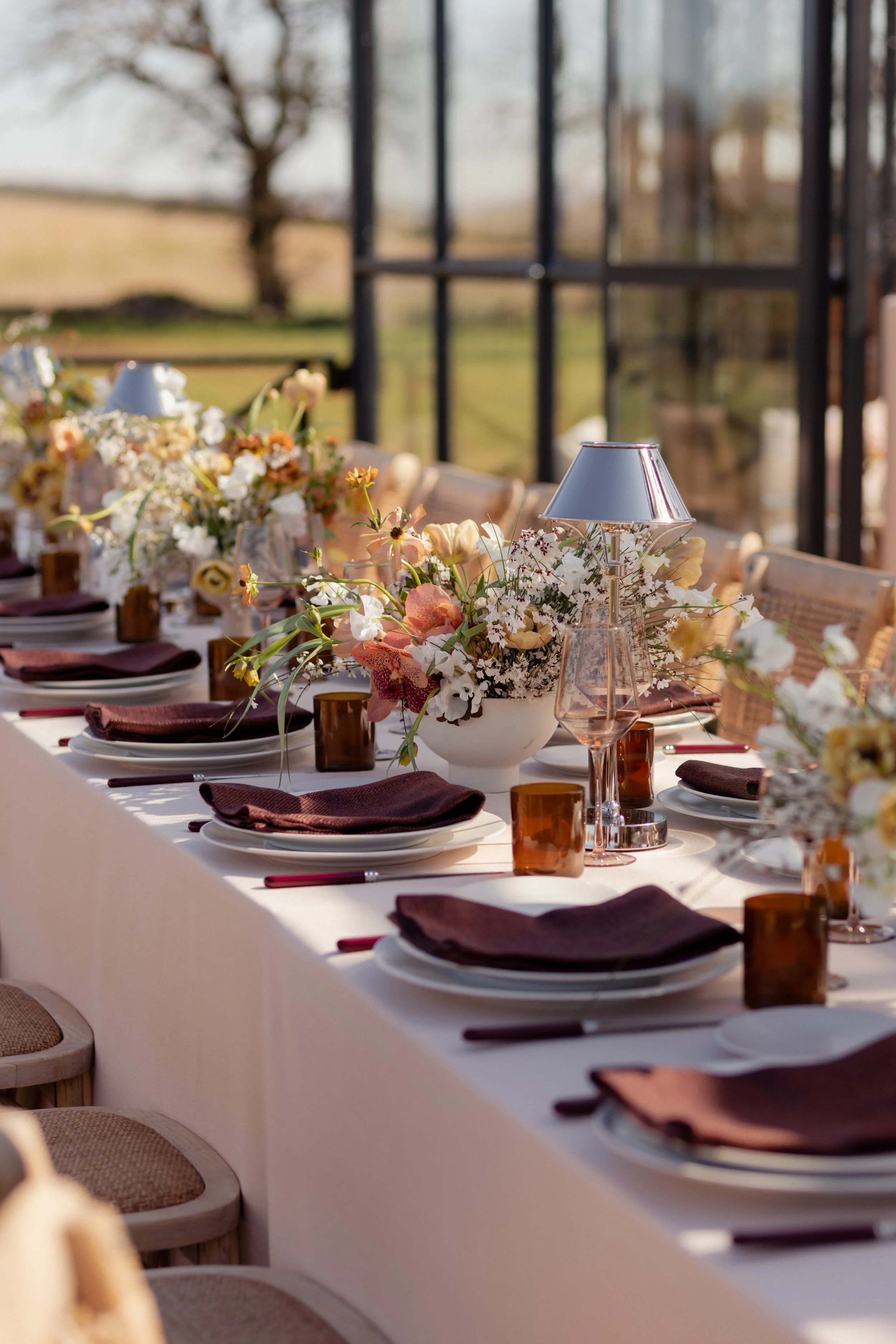A beautifully set outdoor dining table decorated with floral centerpieces, plates, glasses, and lamps, with a scenic view of trees and greenery in the background at The Orangery with Trille Floral.