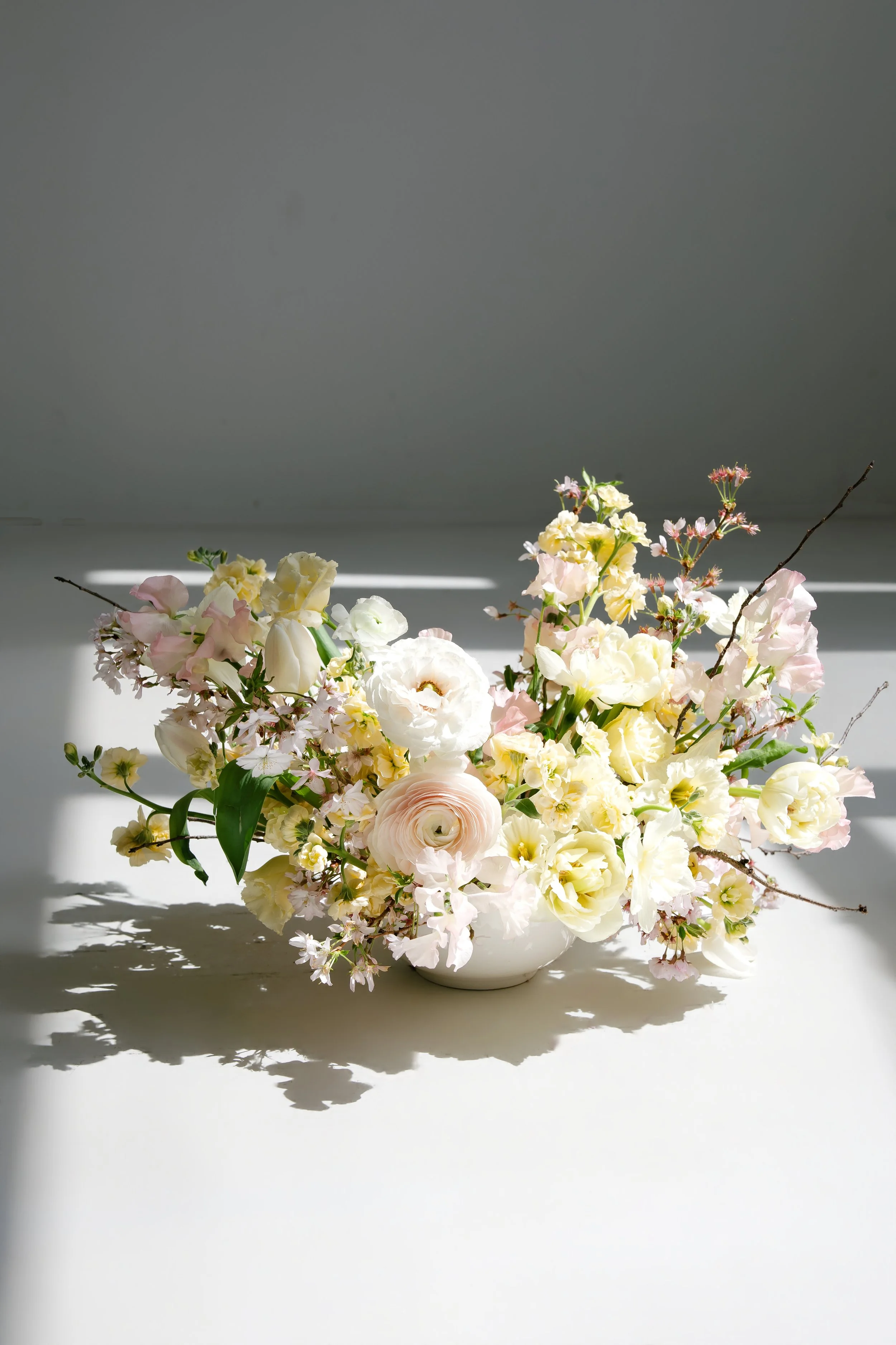 A white ceramic bowl filled with a large bouquet of various white, cream, and light pink flowers, with some pink-tinged blossoms, arranged with green leaves, casting shadows on a white surface and a gray wall background.