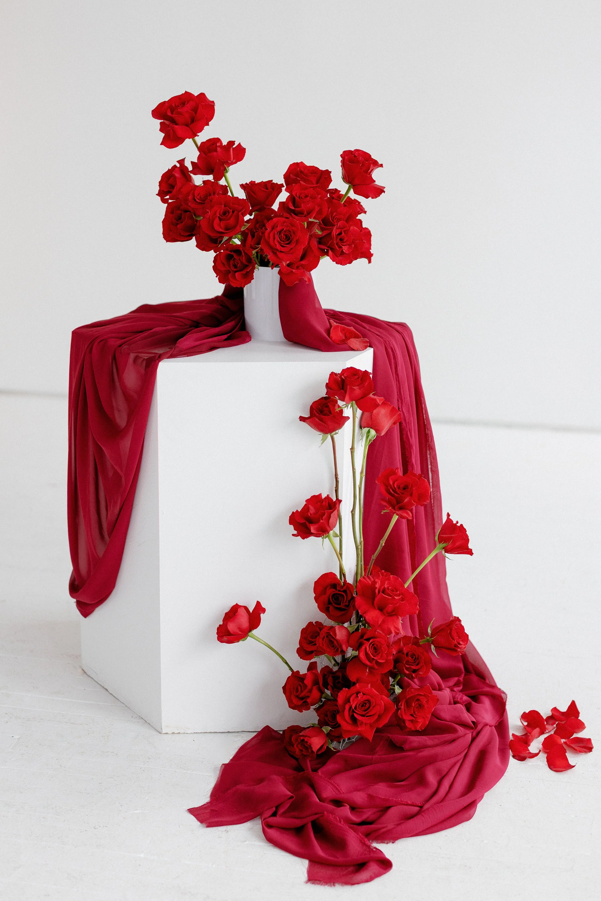 A white vase with red roses on a white pedestal, with red fabric draped around and spilling onto the floor, against a plain white background in Jacalyn Meyvis's photo studio.