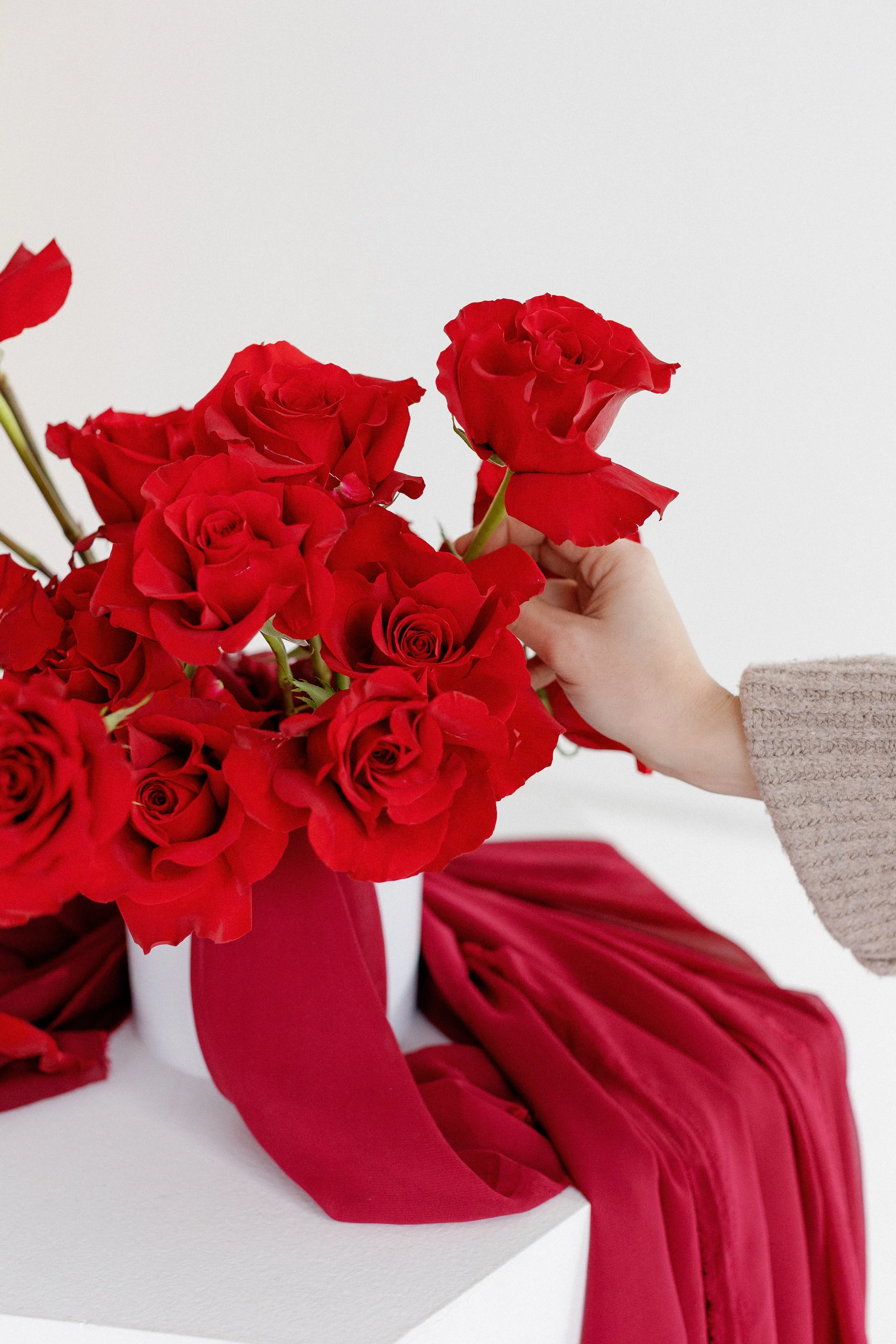 Person holding a red rose in front of a vase of red roses, with red ribbon and fabric draped underneath, against a plain white background.