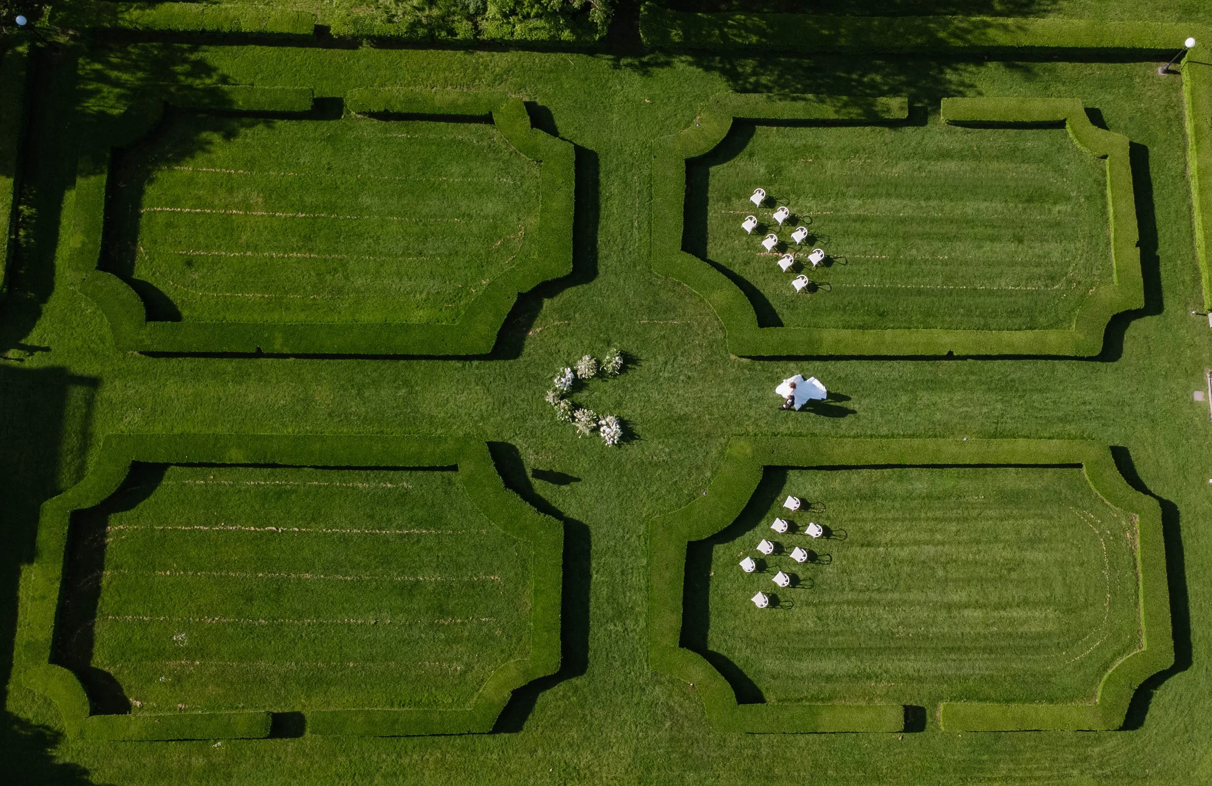 A top-down aerial view of a neatly trimmed hedge maze with a person dressed in white at the center. White chairs are arranged in a semi-circle nearby, and trees cast shadows on the green grass with a huge ground floral ceremony at Geneva on the Lake