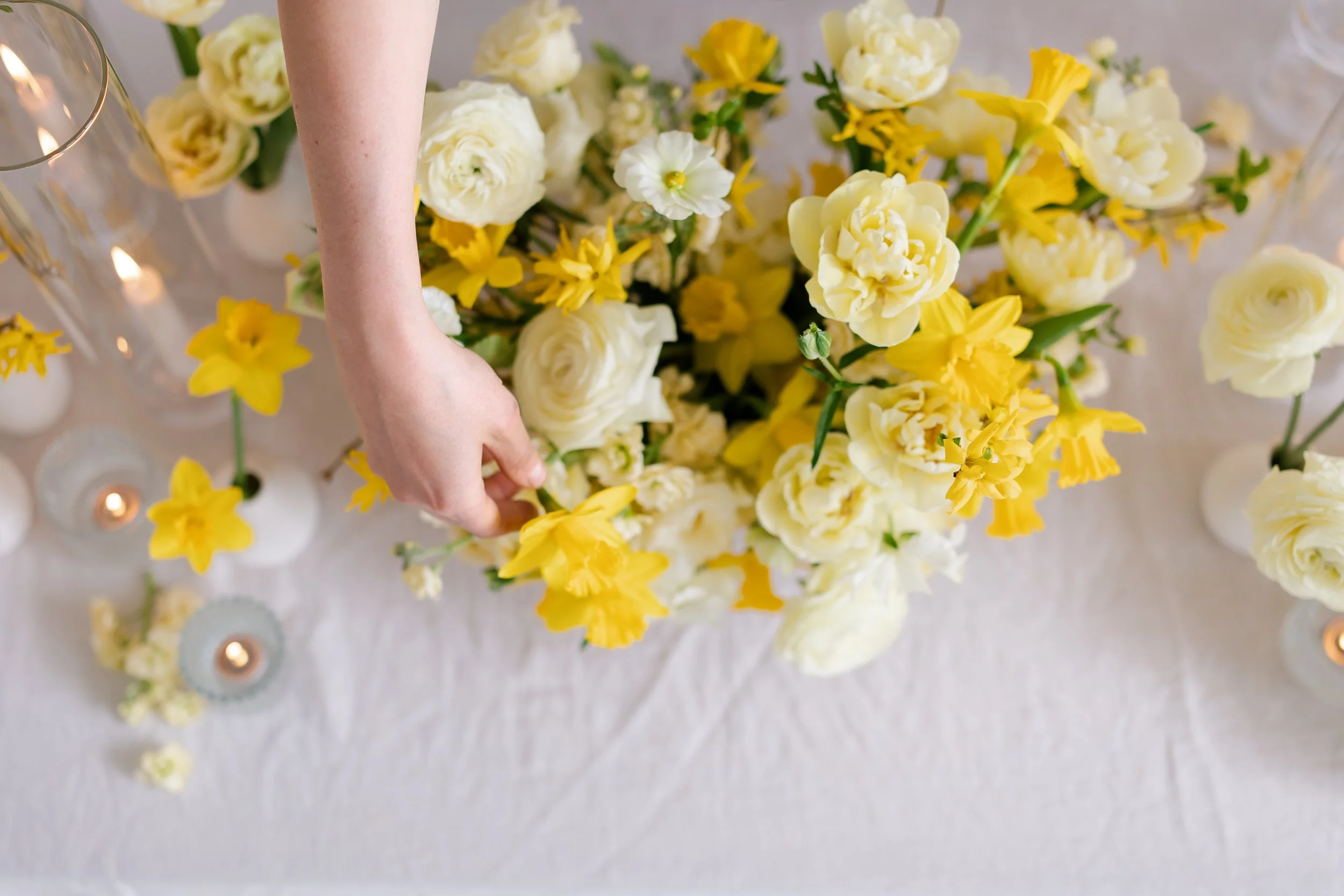 Close-up of a person's arm reaching toward a bouquet of white and yellow flowers, with lit candles in glass holders and scattered flower petals on a white surface.