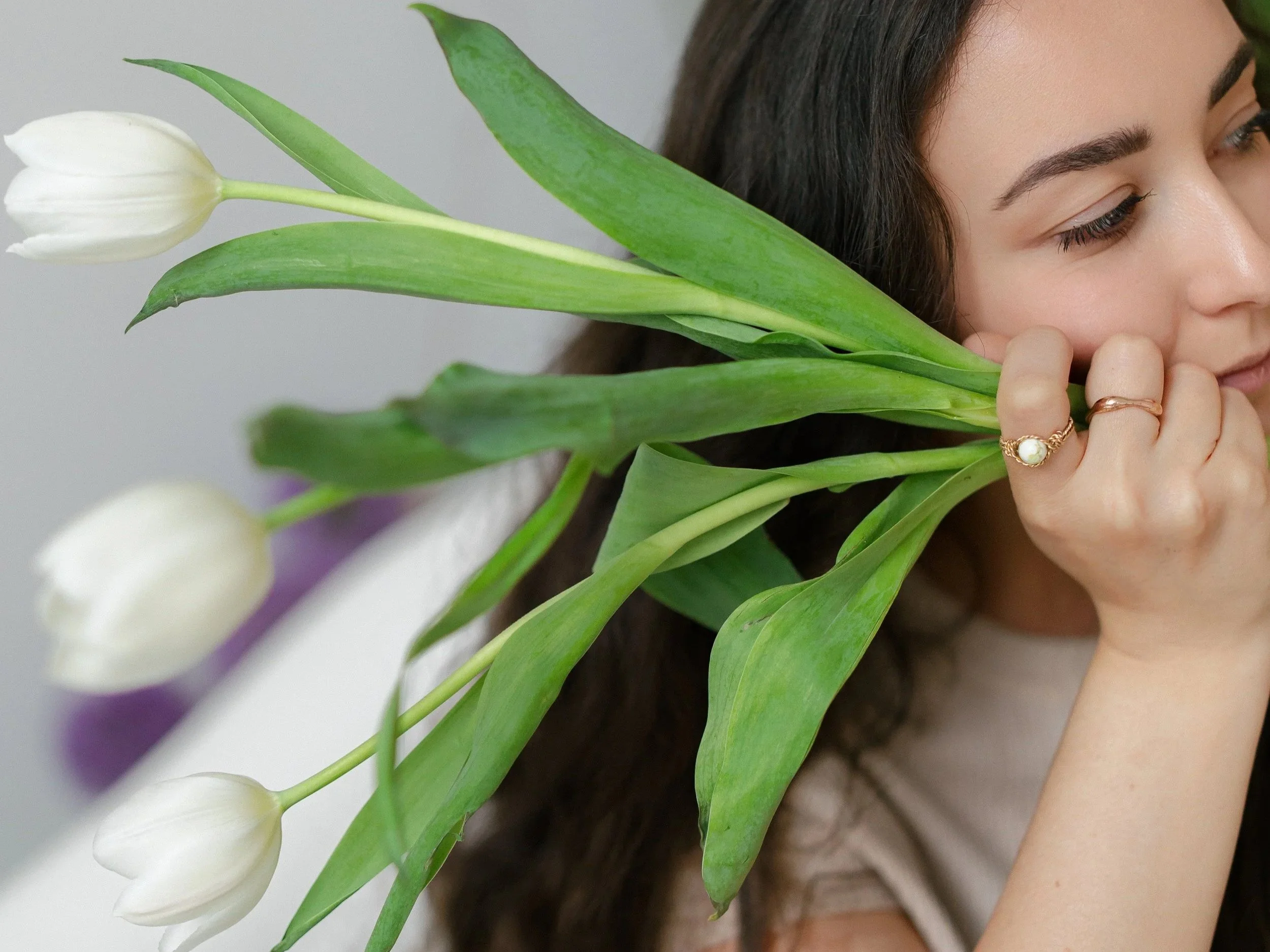 A young woman with dark hair, makeup, and rings holds white tulips close to her face against a light gray background.