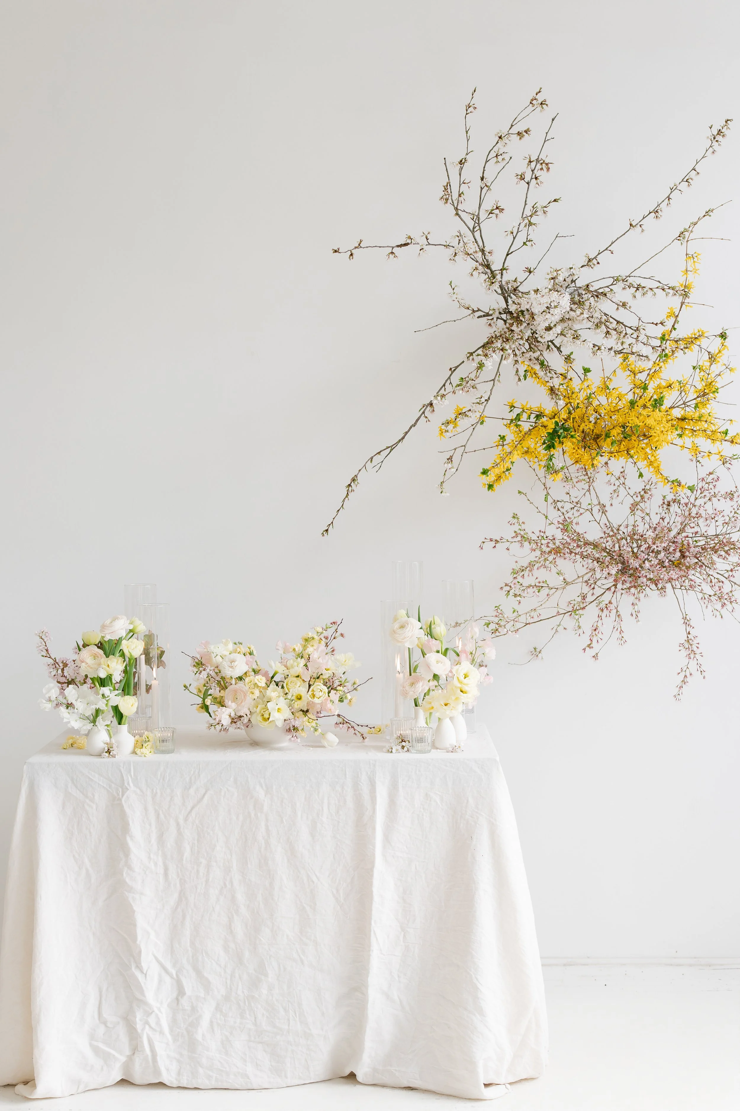 A white tablecloth-covered table decorated with various white, cream, and pink flowers in vases, with a tall branch of pink blossoms and yellow flowers attached to the white wall behind created by Midnight Moss Co.