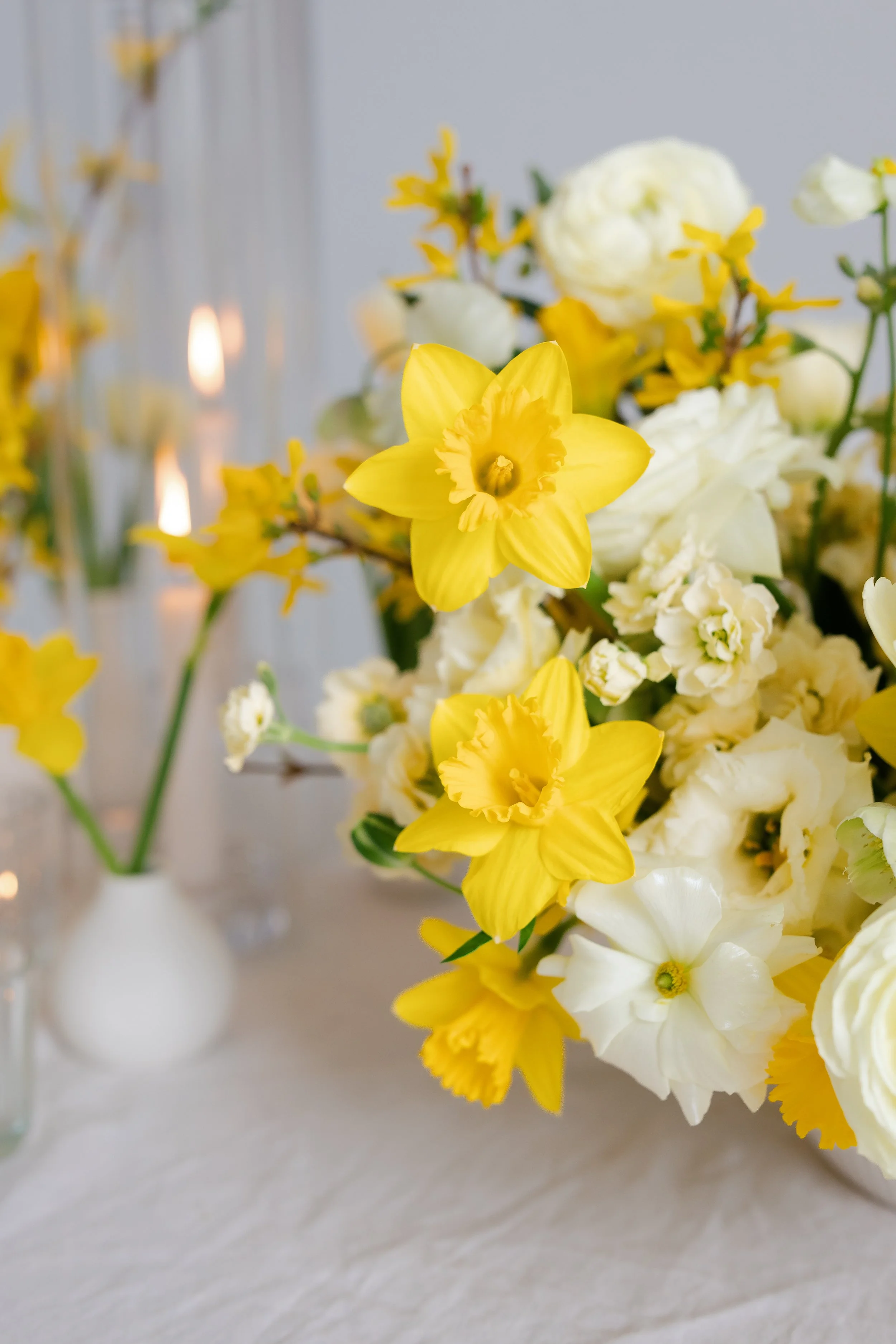A bouquet of yellow and white flowers in a white vase on a white surface, with a blurred background.