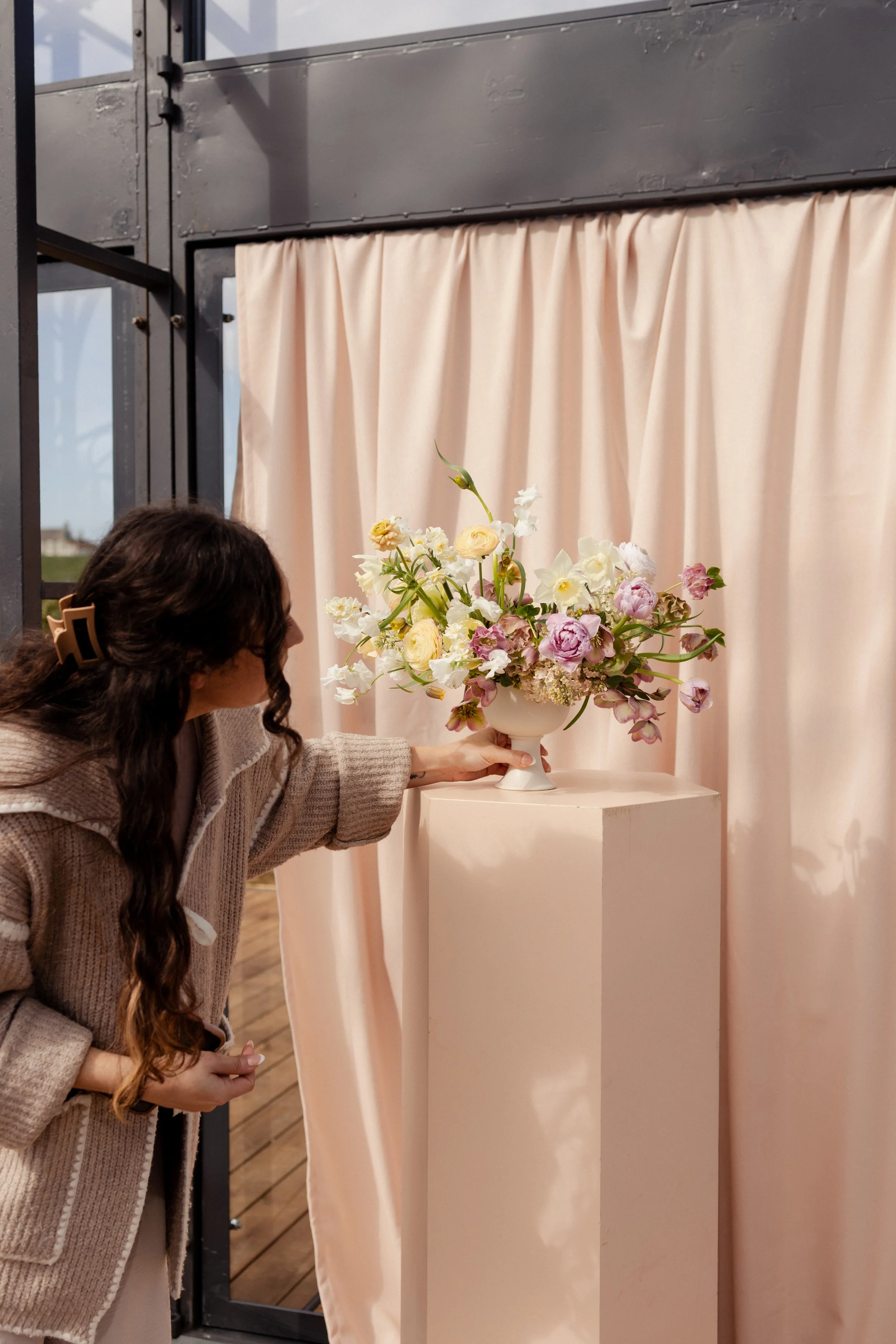 A woman with long curly hair, wearing glasses, a beige sweater is placing flowers in a white Gaia vessel on a pedestal. The bouquet contains white stock, pink sweet pea and lilacs. The background has beige curtains and a black metal structure.