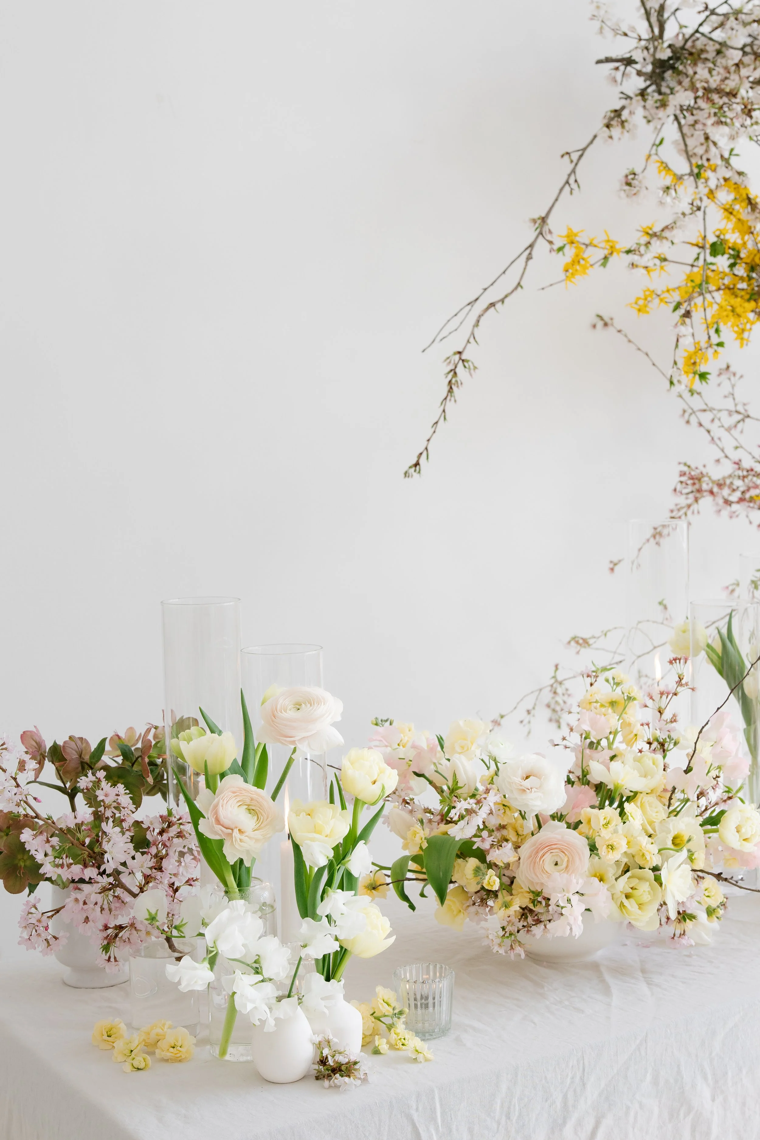 A floral arrangement with white, pink, and yellow flowers in various vases on a white table against a white background, with some branches hanging from the top right corner.