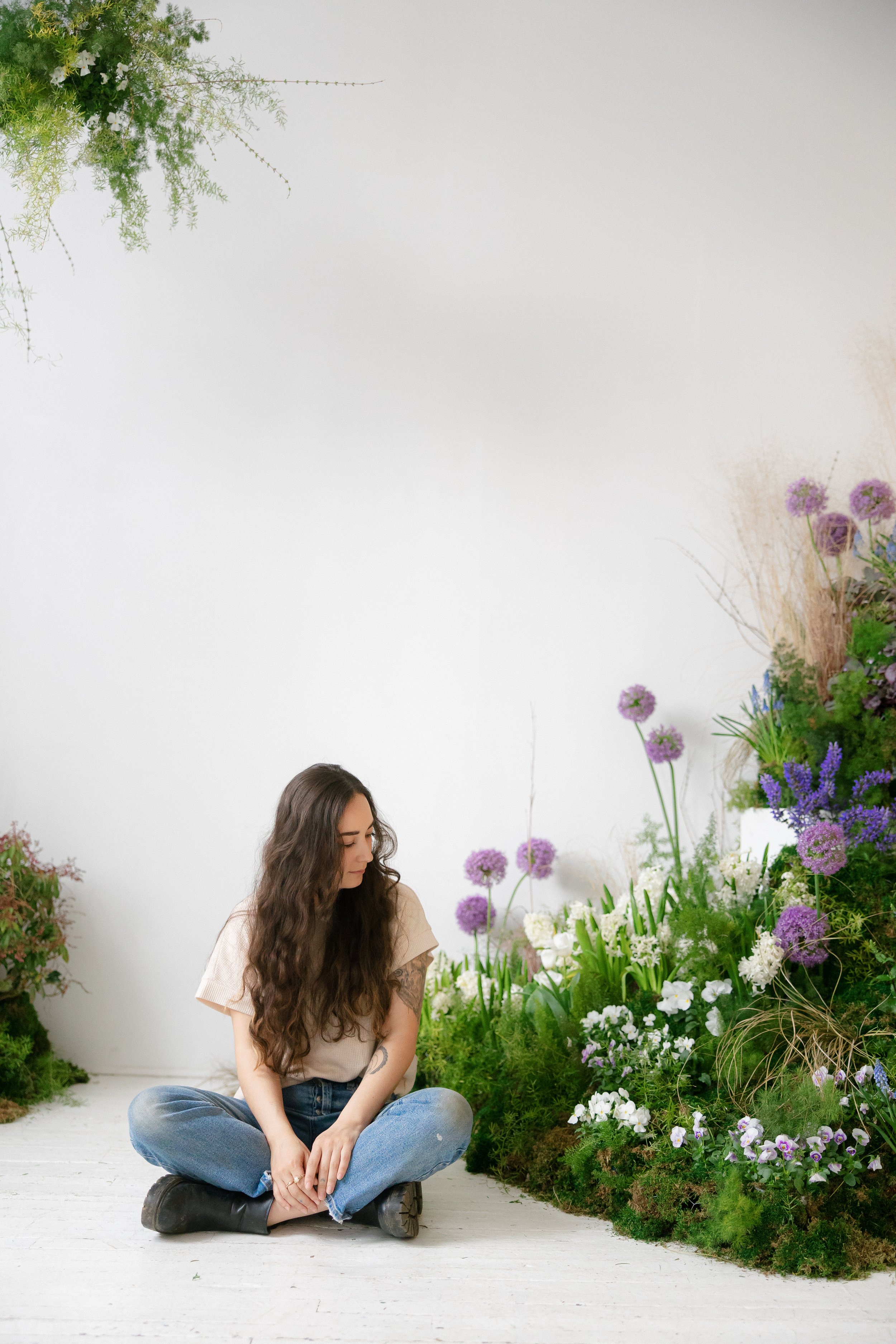 A woman with long brown hair, wearing a beige top and blue jeans, sitting cross-legged on a white wooden floor in front of a large, lush flower and greenery display, looking down with a gentle smile.