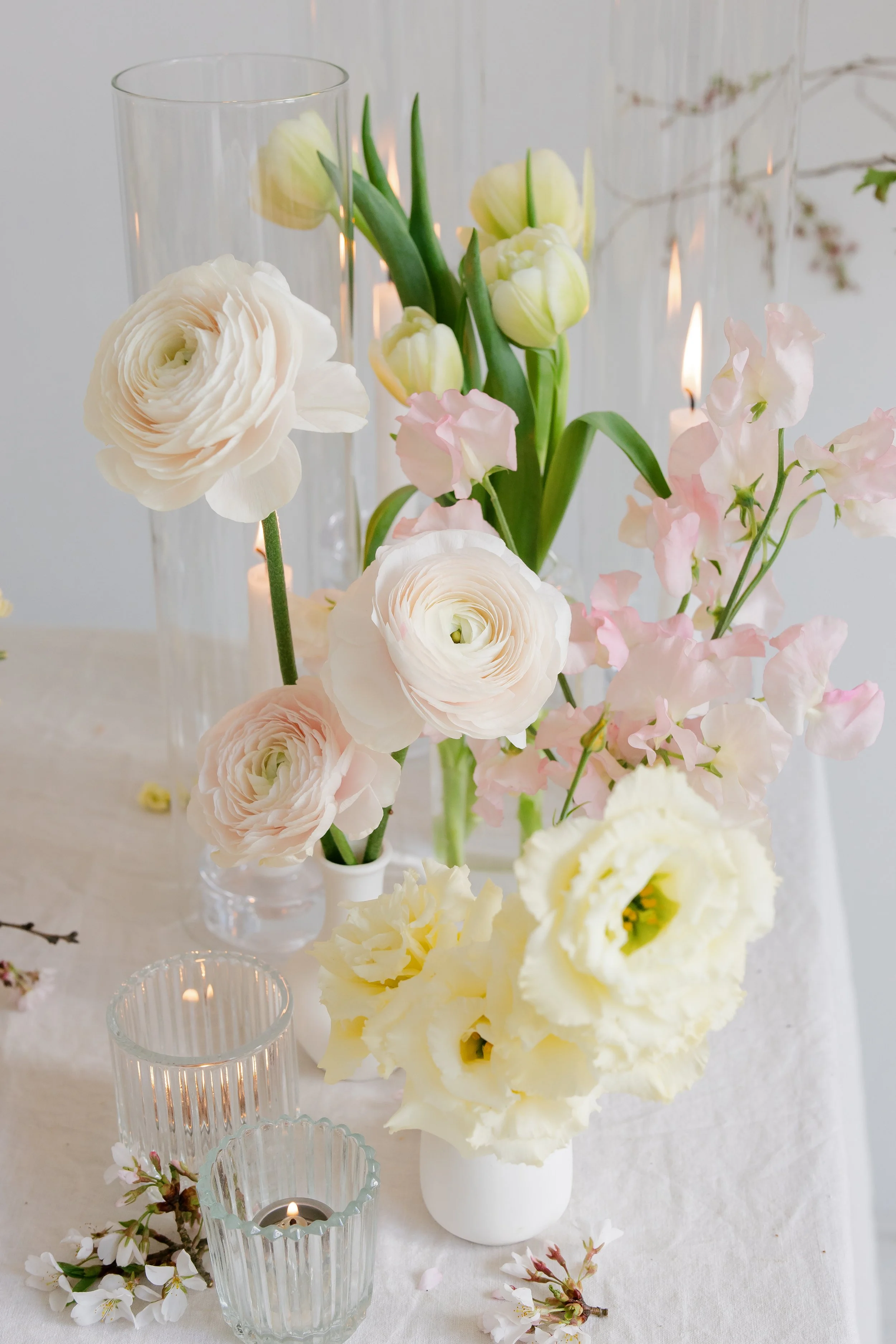 A floral arrangement consisting of white and pale pink flowers, including ranunculus, tulips, and delphiniums, in glass vases with tea light candles placed nearby on a white tablecloth in Syracuse, NY.