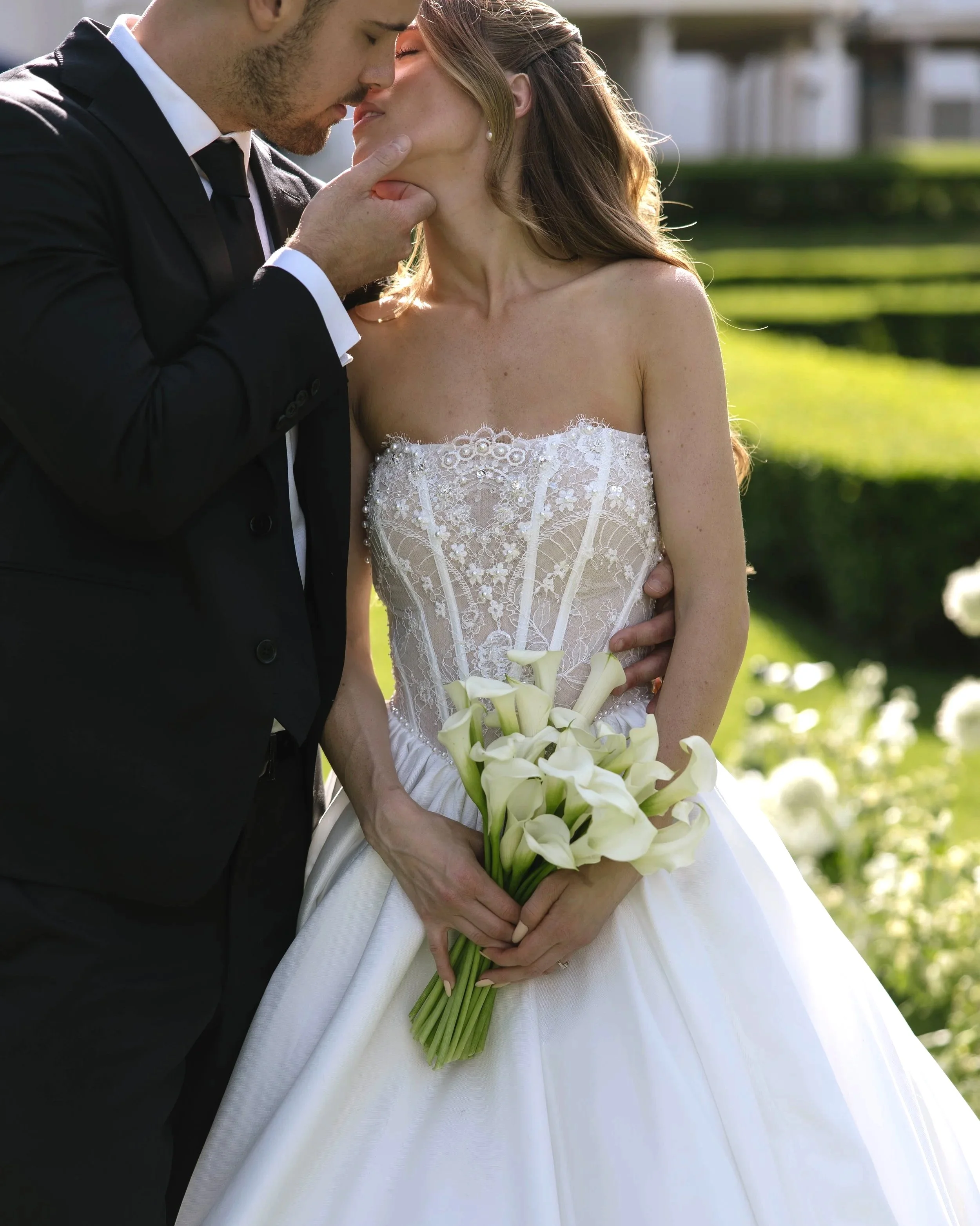 A couple shares a kiss in the midst of their wedding ceremony at Geneva on the Lake, the groom gently holding the bride's chin while she holds a bouquet of white calla lilies, submersed in a sea of white alliums, calla lilies, hydrangea, amaranthus.
