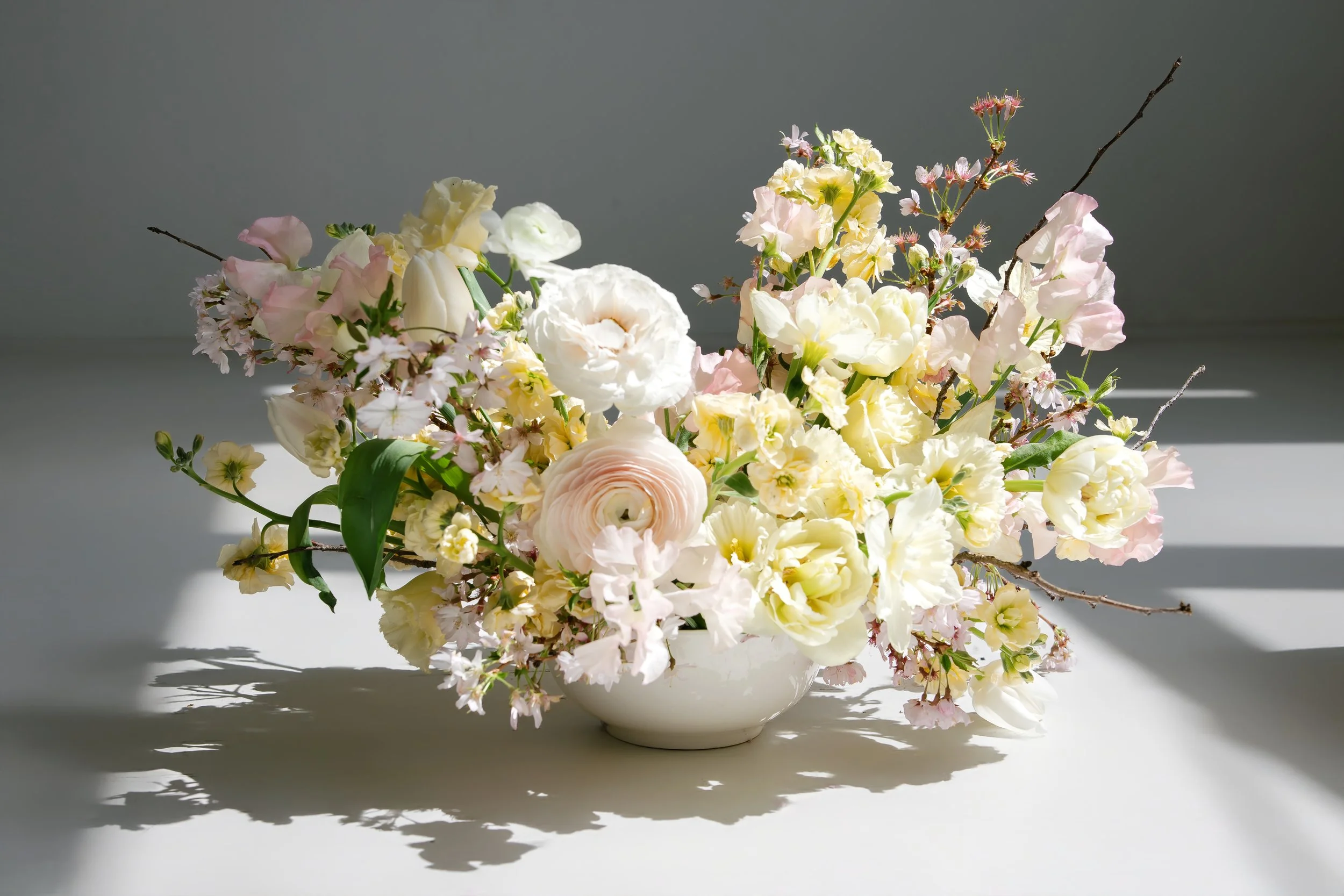 A white ceramic vase filled with a bouquet of pale yellow, pink, and white flowers with green leaves, placed on a simple white surface with shadows cast underneath.