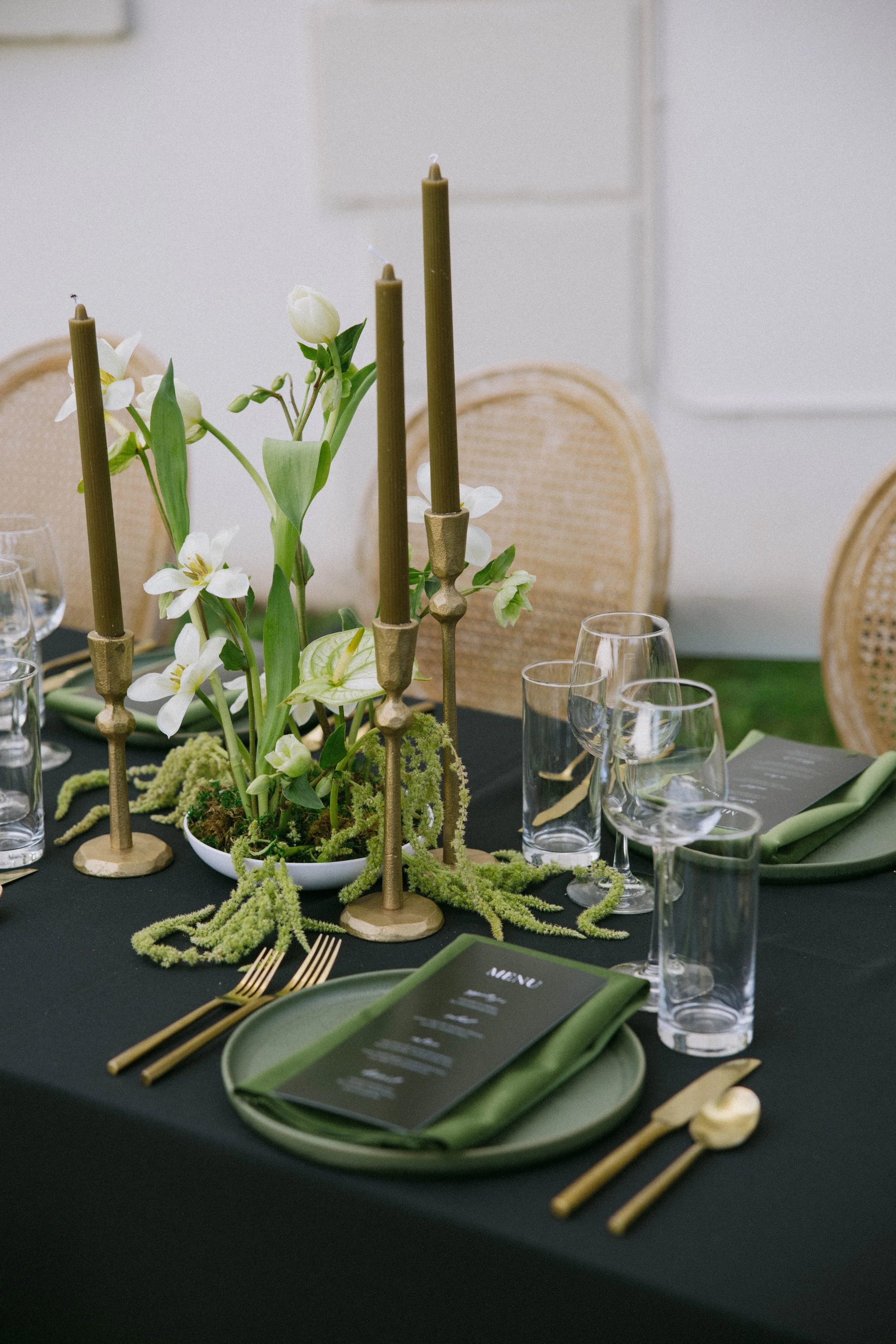 Elegant dining table with black tablecloth, green and white ikebana florals, gold candlesticks, glassware, green napkins, black menus, gold cutlery, and neutral-colored chairs in the background for a wedding reception in Geneva, NY.