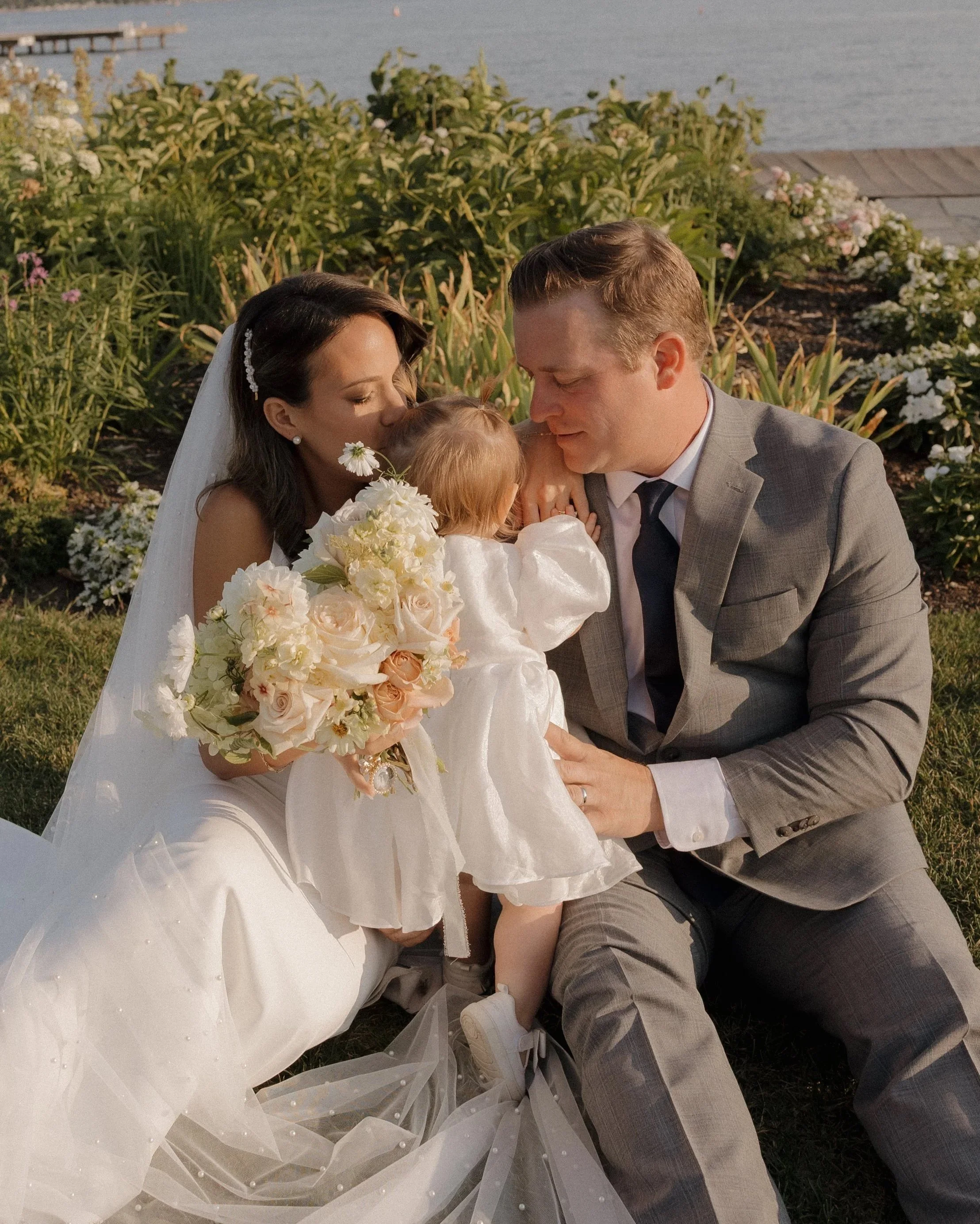 A bride and groom kneel by a young girl during an outdoor wedding ceremony by the water at The Lake House in Canandaigua, with greenery and flowers in the background.