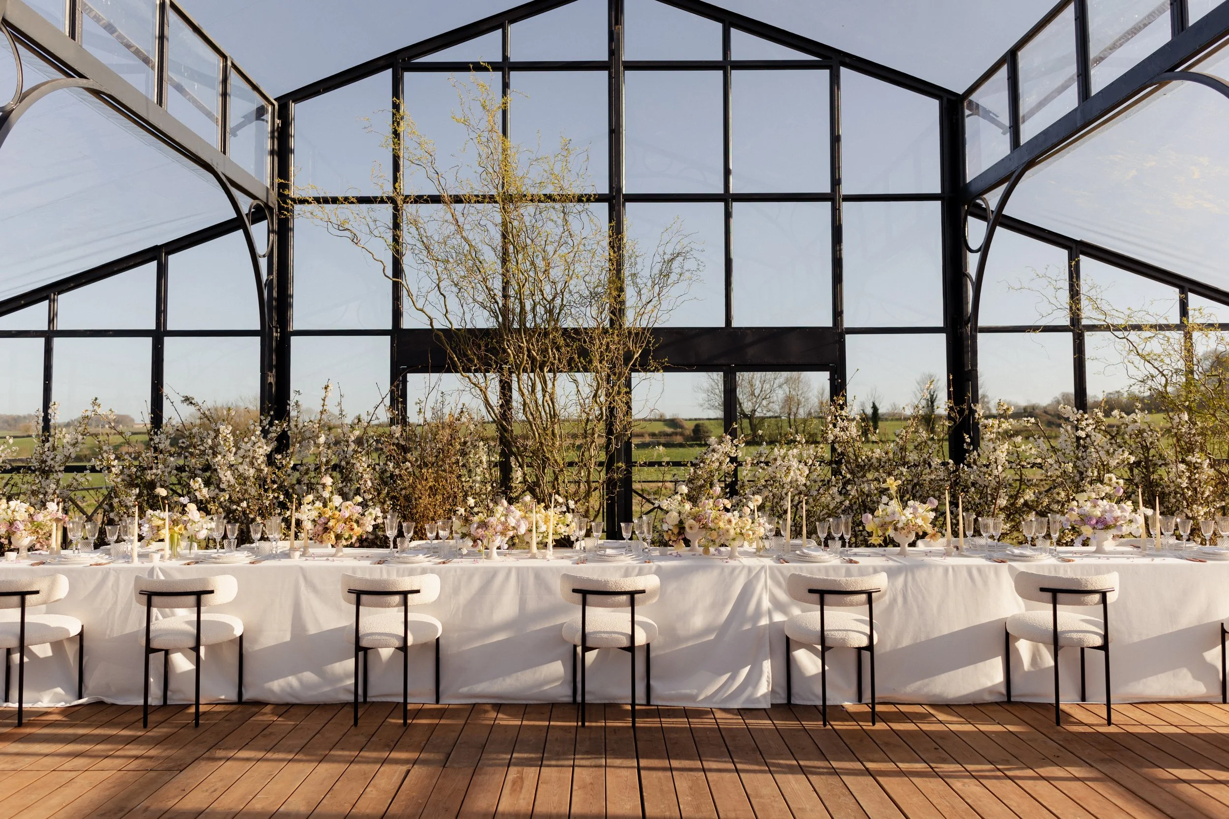 Decorated banquet table set with floral arrangements, white tablecloth, glassware, and candles inside a geometric glass building, with trees and a field visible outside for a corporate function in Bath, England.