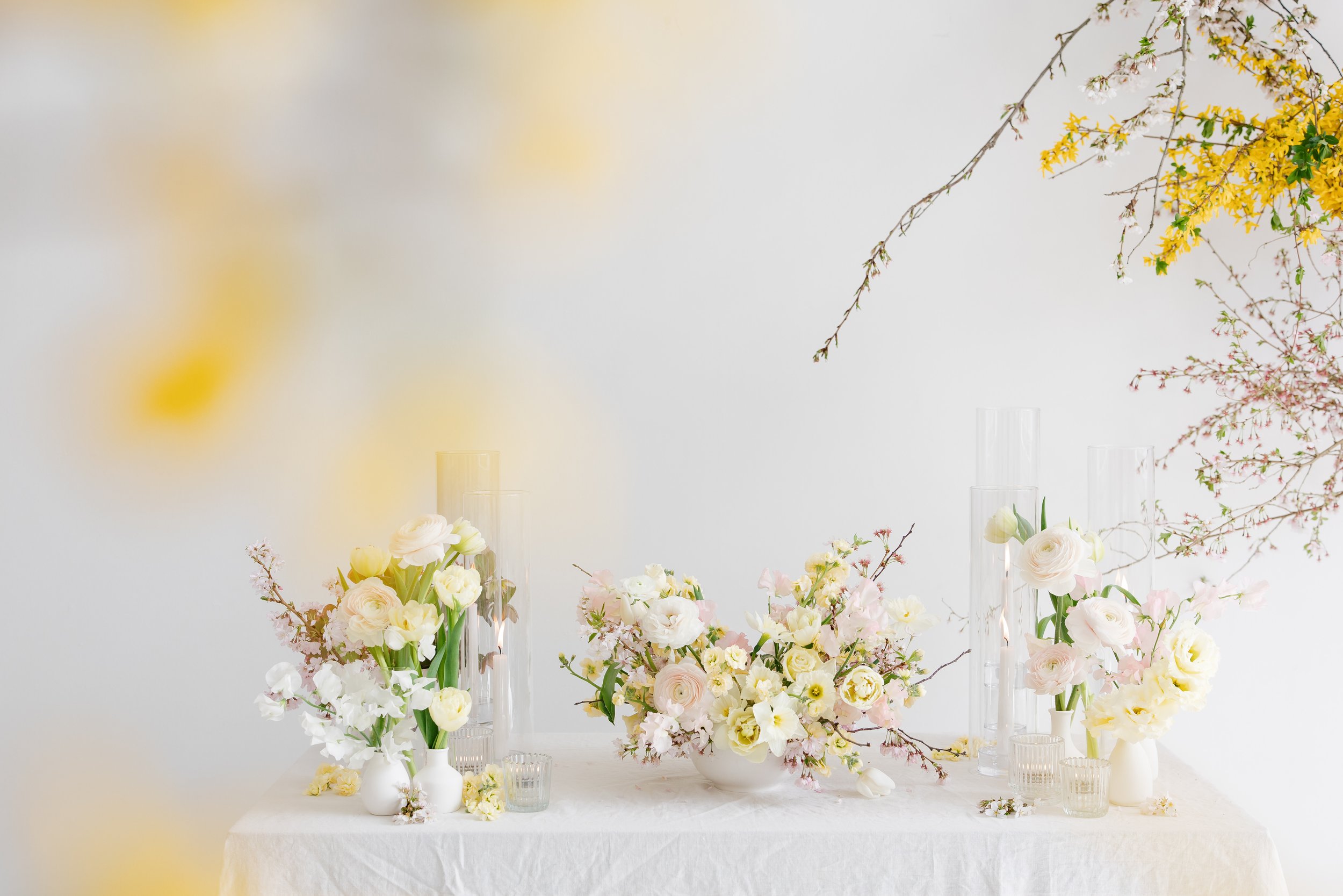 Elegant floral arrangement with white and blush flowers, green foliage, and pink cherry blossoms in clear glass vases on a white tablecloth against a simple white background.