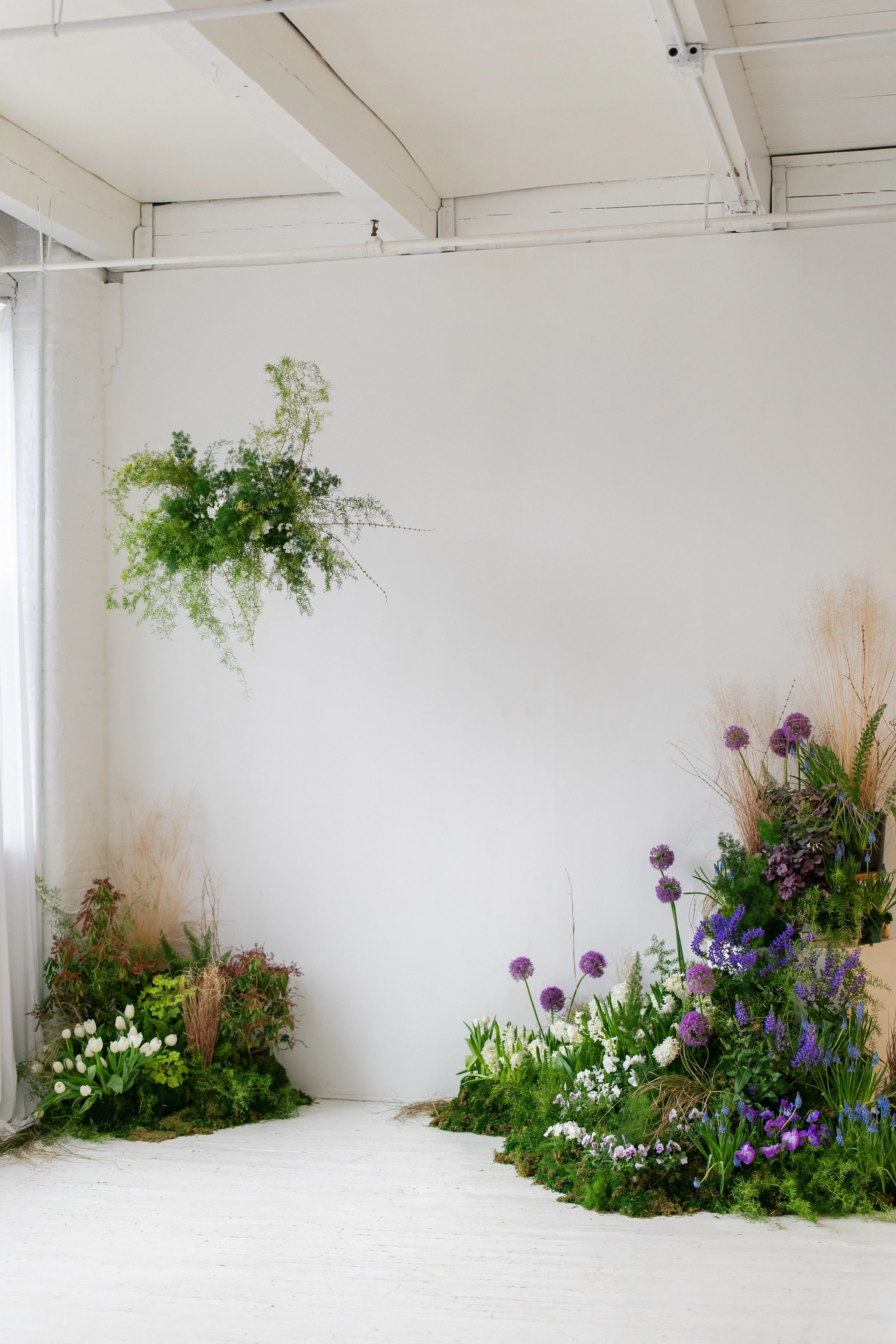 An indoor floral display in Lindsay Stephany Photographer's studio at the Artist's Alley in Rochester, NY, featuring a variety of flowers and plants arranged on the floor and some hanging from the wall by Midnight Moss Co.