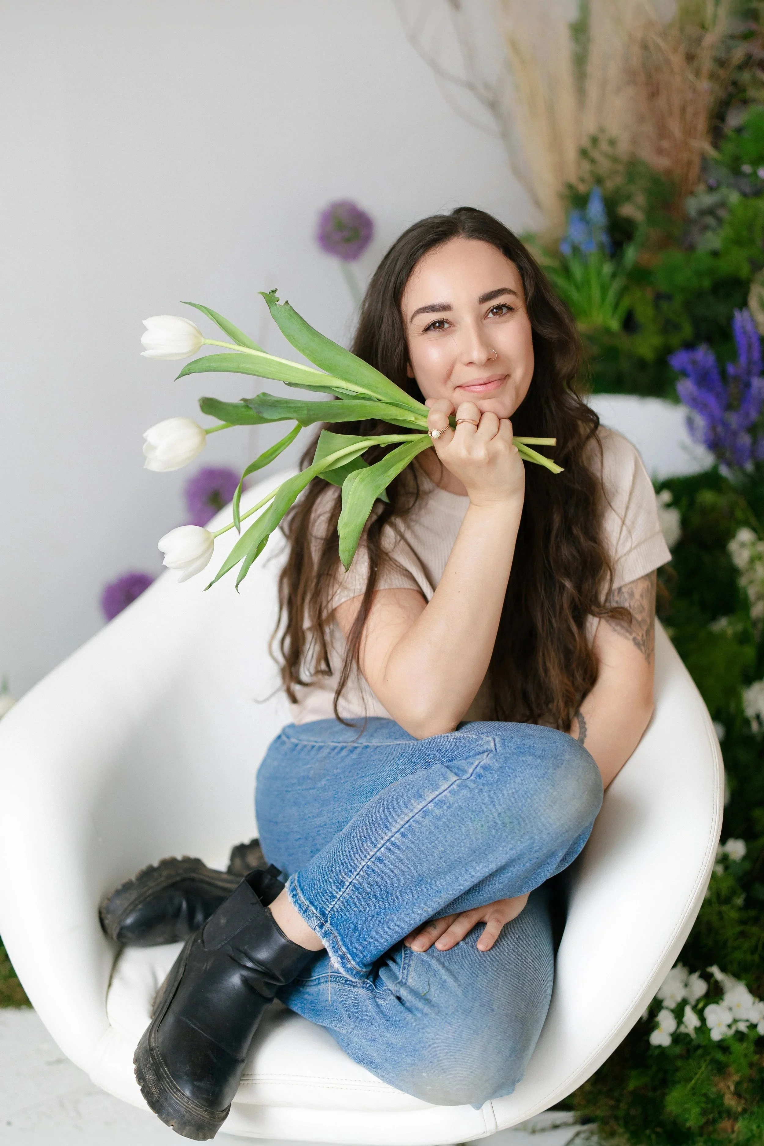 A young woman with long curly dark hair sitting in a white modern chair, holding a bunch of white tulips near her face, smiling softly, with a backdrop of purple alliums and green bear grass and foliage at the Anderson Arts Building.