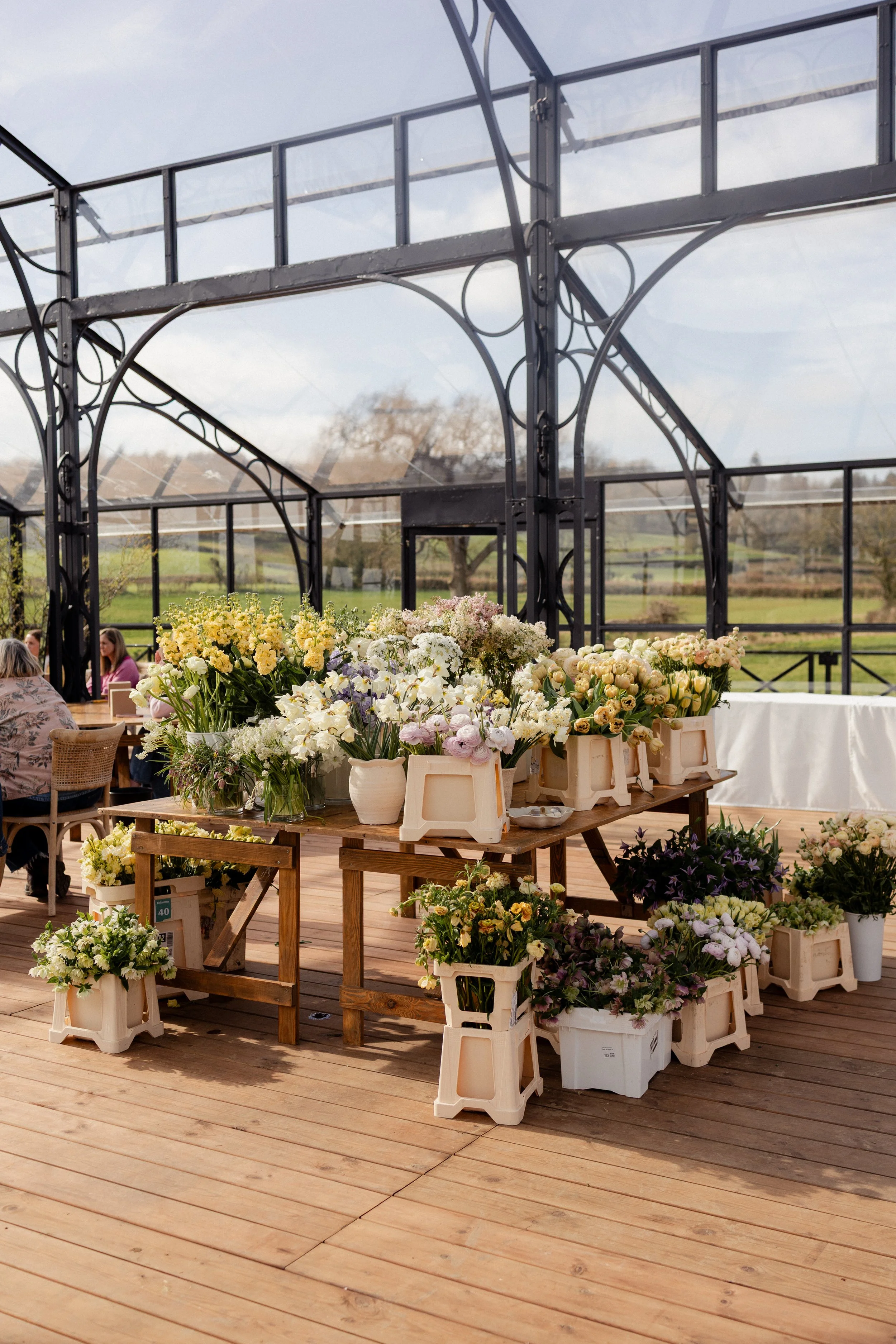 Display of various flowers arranged in baskets and containers at a flower market or event, set outdoors under a greenhouse structure for a wedding ceremony.