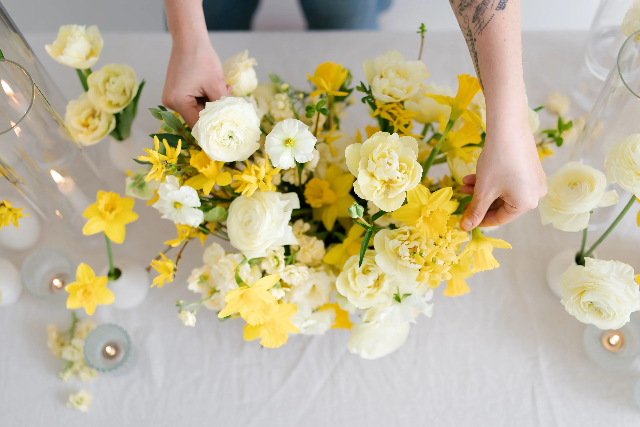 Hands arranging a bouquet of yellow and white flowers on a table with candles.