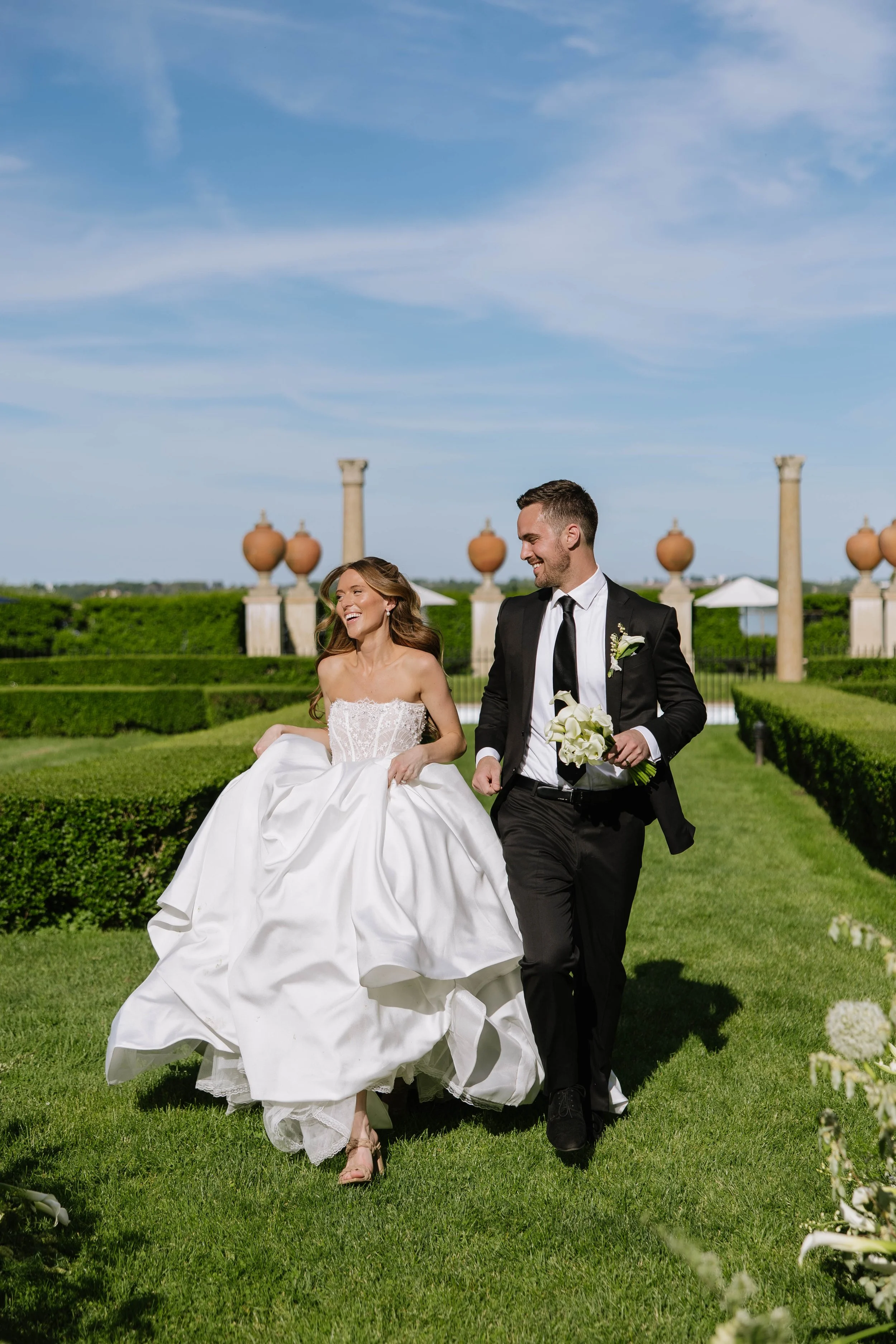 A bride and groom running together outdoors in a garden on a sunny day, with the bride smiling and holding her dress, bouquet of calla lilies and the groom smiling at Geneva on the Lake.