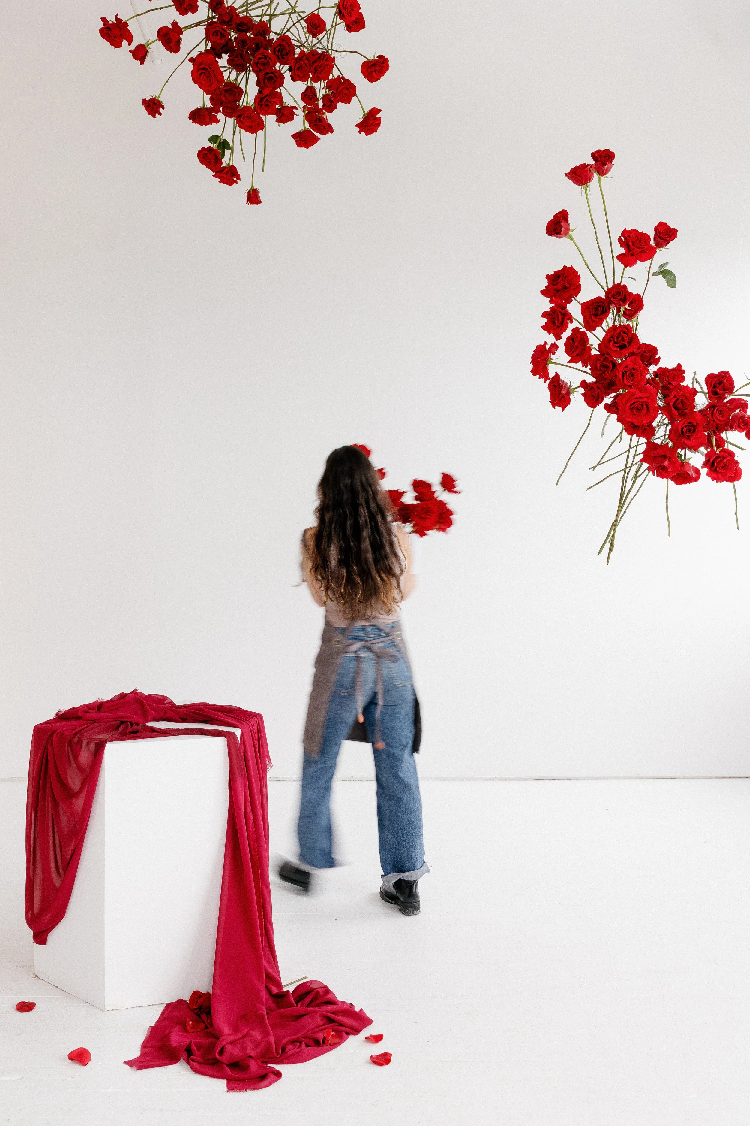 Woman with long curly hair in jeans and a light top working in an art gallery with red roses and fabric art installations on the walls and a pedestal with red fabric draped over it.