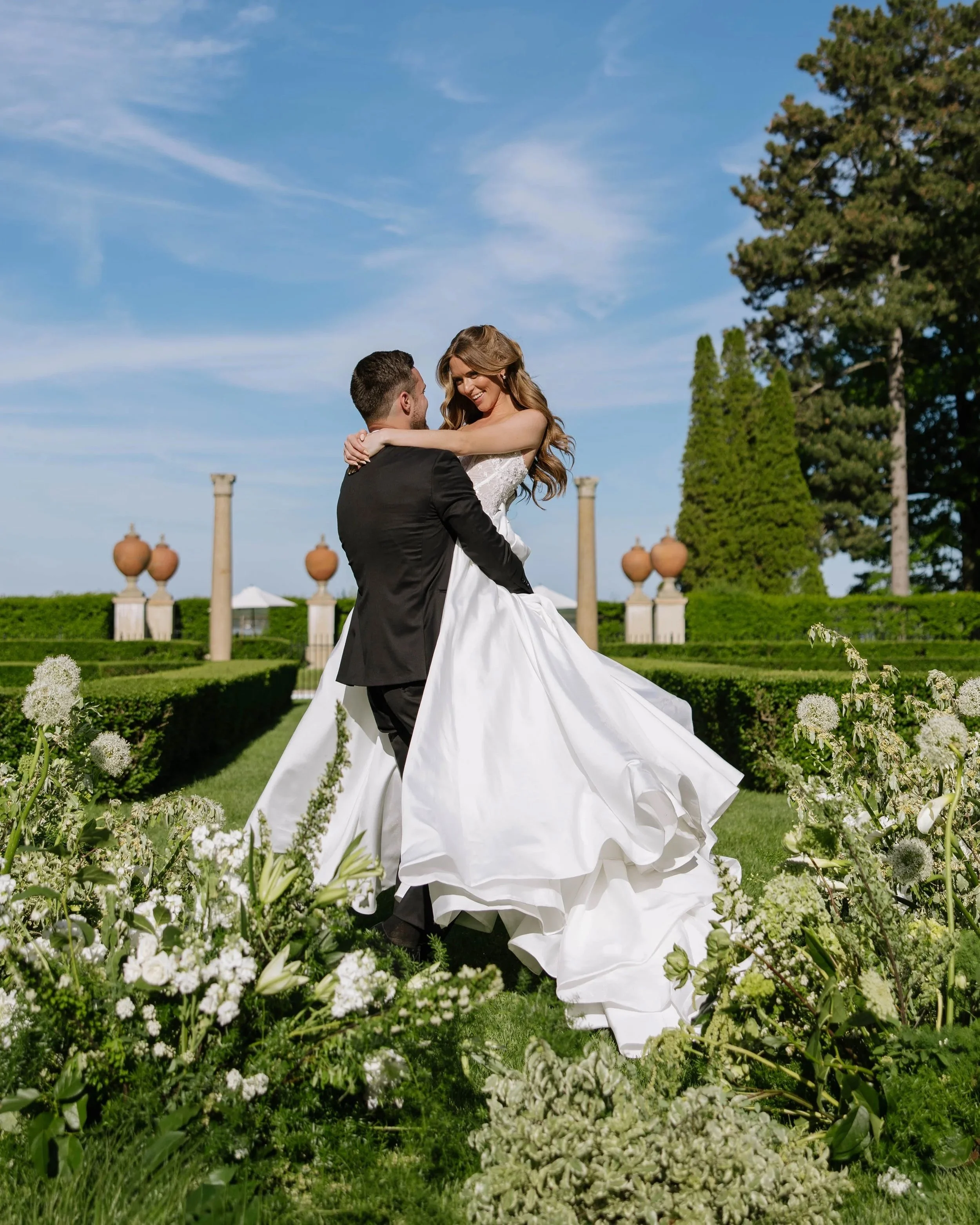 A man in a black suit and a woman in a white wedding dress are dancing outside in a garden with green grass and white flowers, under a clear blue sky with a few clouds.