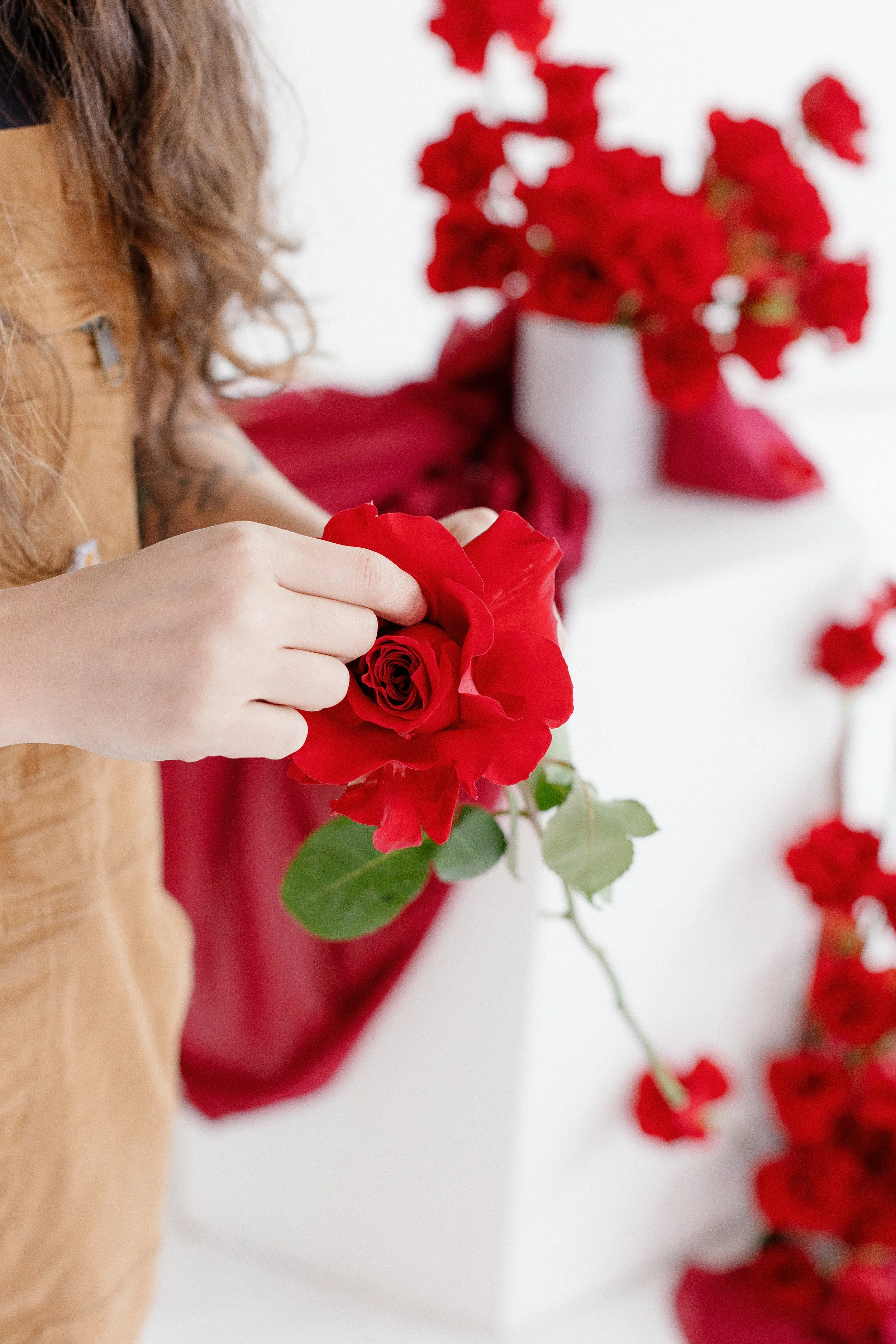 A person with curly hair and a tattoo on their arm arranging a red rose. In the background, there is a white vase with red flowers and red fabric. The image is bright with a minimalistic style.