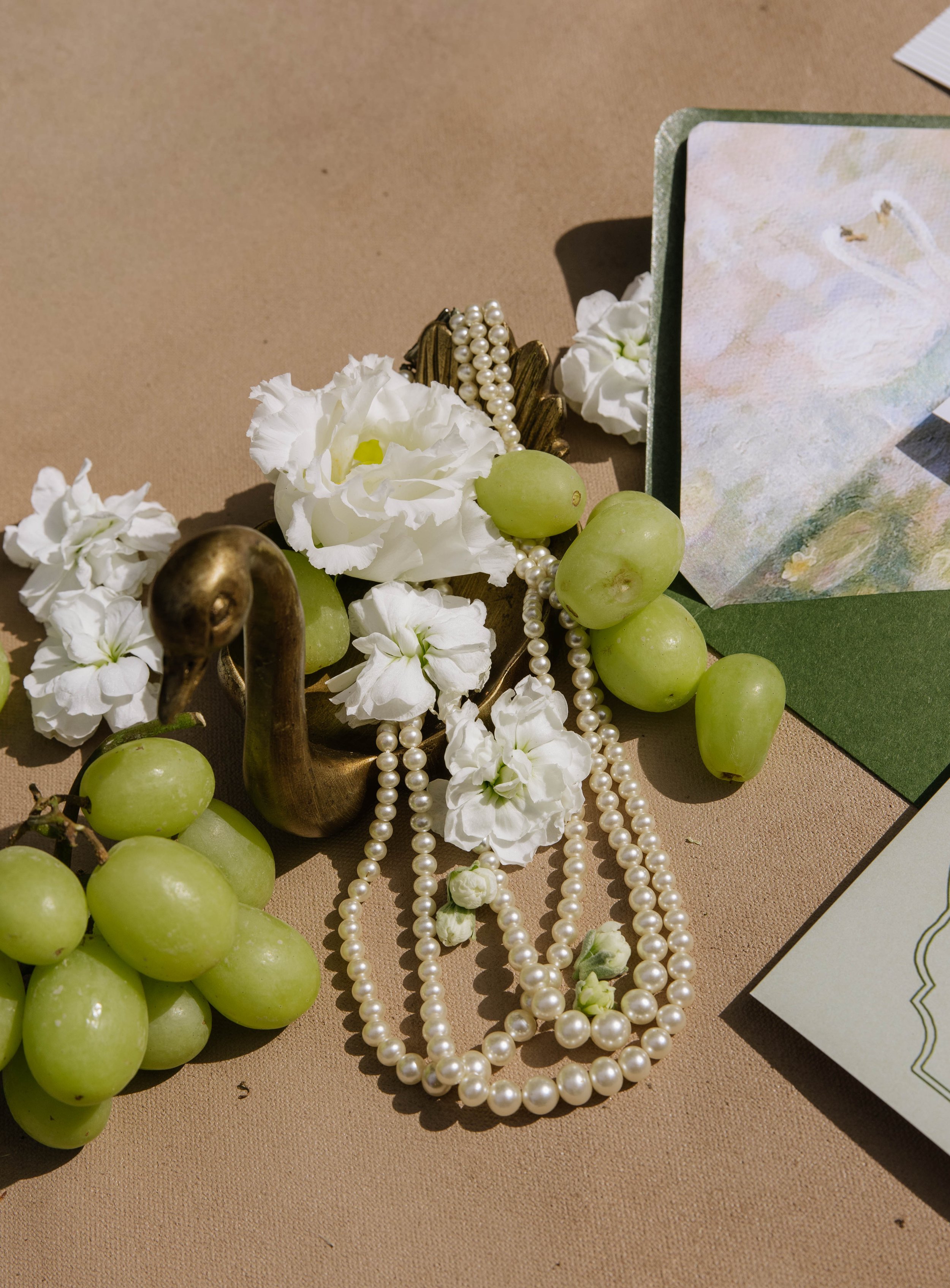 A still life arrangement of green grapes, white stockflowers, a pearl necklace, a brass swan figurine, and a painted card on a beige surface at Geneva on the Lake.