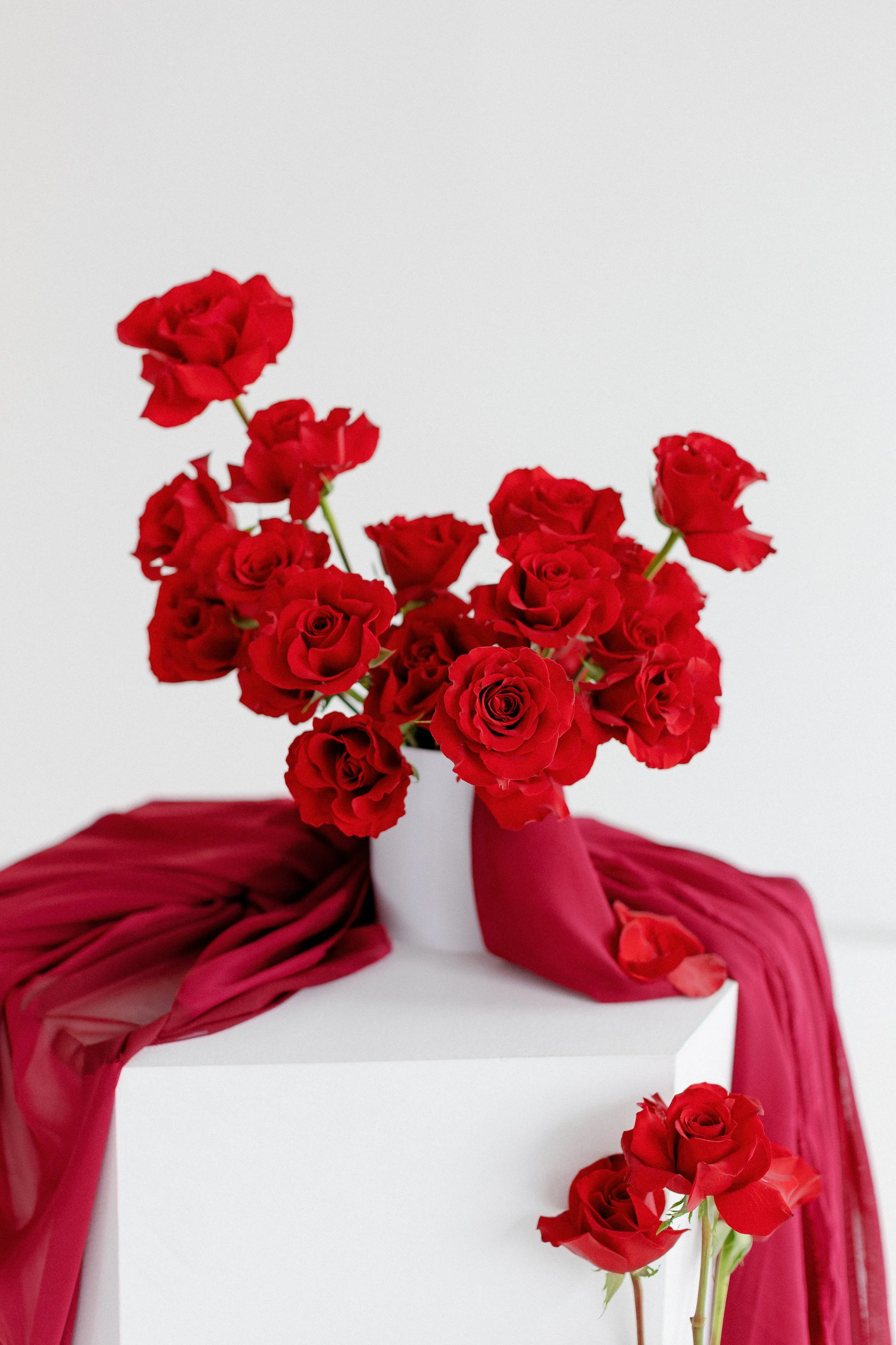 A bouquet of red roses arranged in a white vase, with red fabric draped around the white surface, and some petals scattered on the surface, against a plain light background.