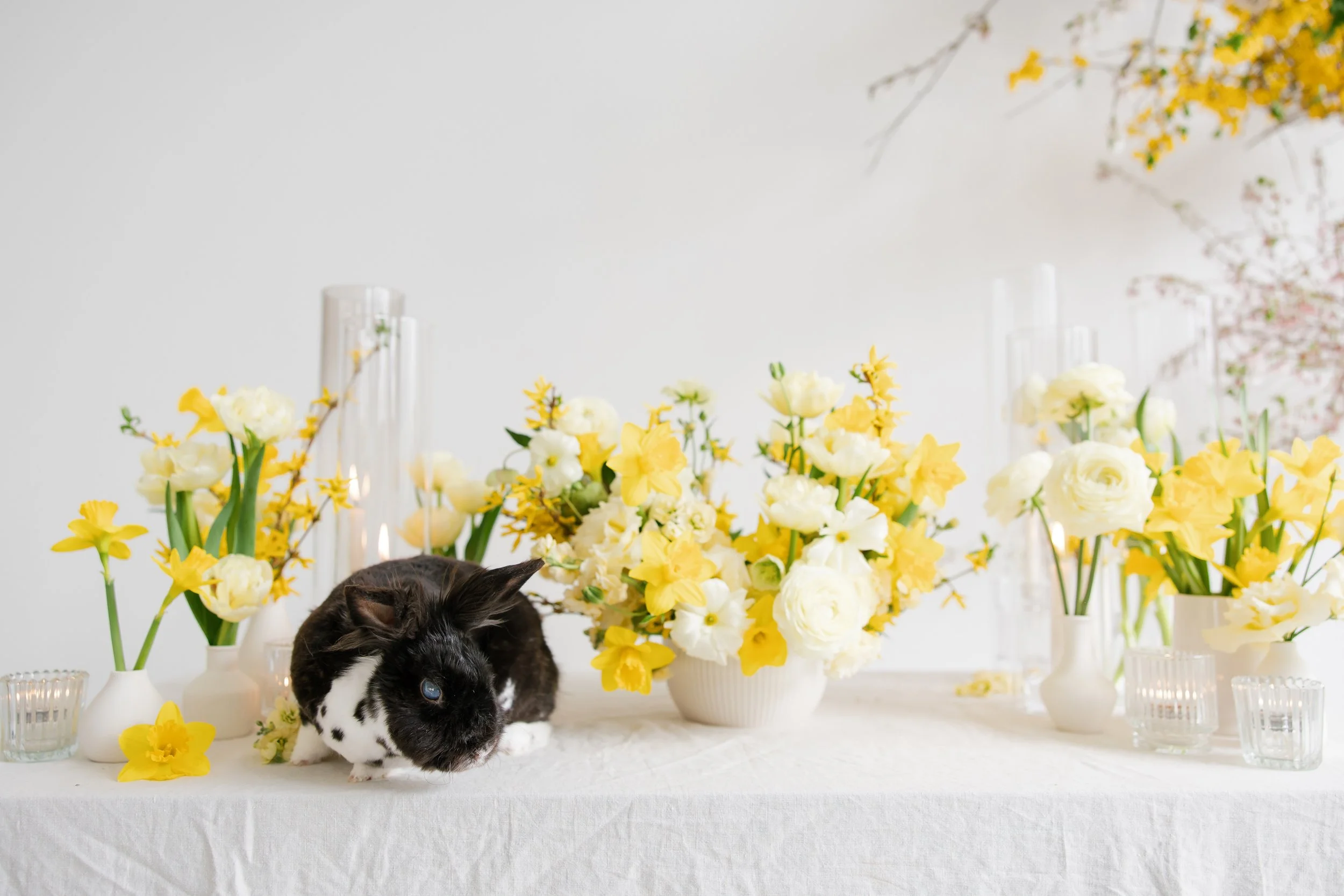 A table decorated with yellow and white flowers, with clear glass candleholders, and a black and white rabbit sitting among the flowers for a wedding reception in Rochester, ny.
