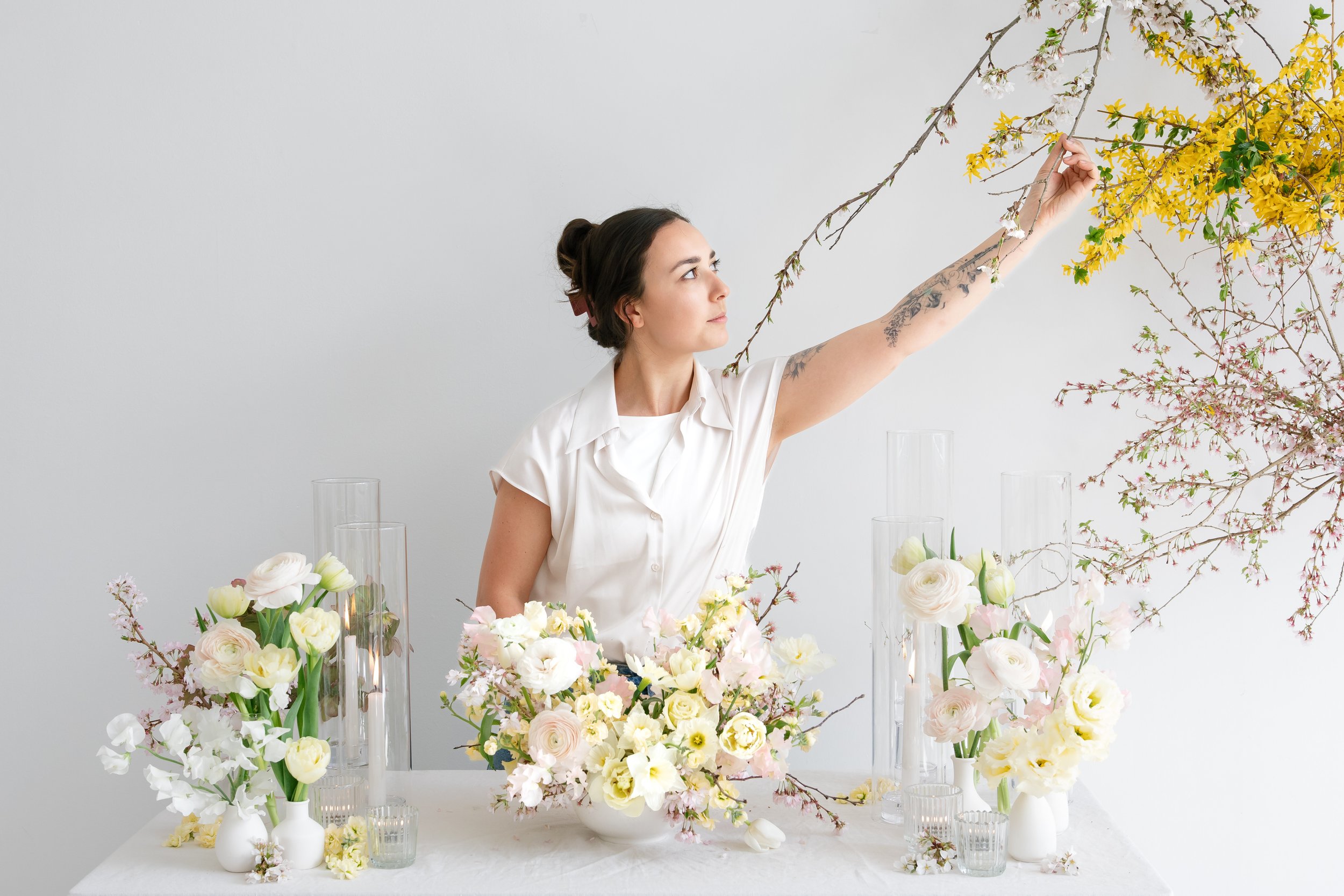 A woman with dark hair in a bun and floral tattoos on her arm arranging pink and white flowers on a white table, surrounded by various vases of flowers and branches, set in The Lake House in Canandaigua.
