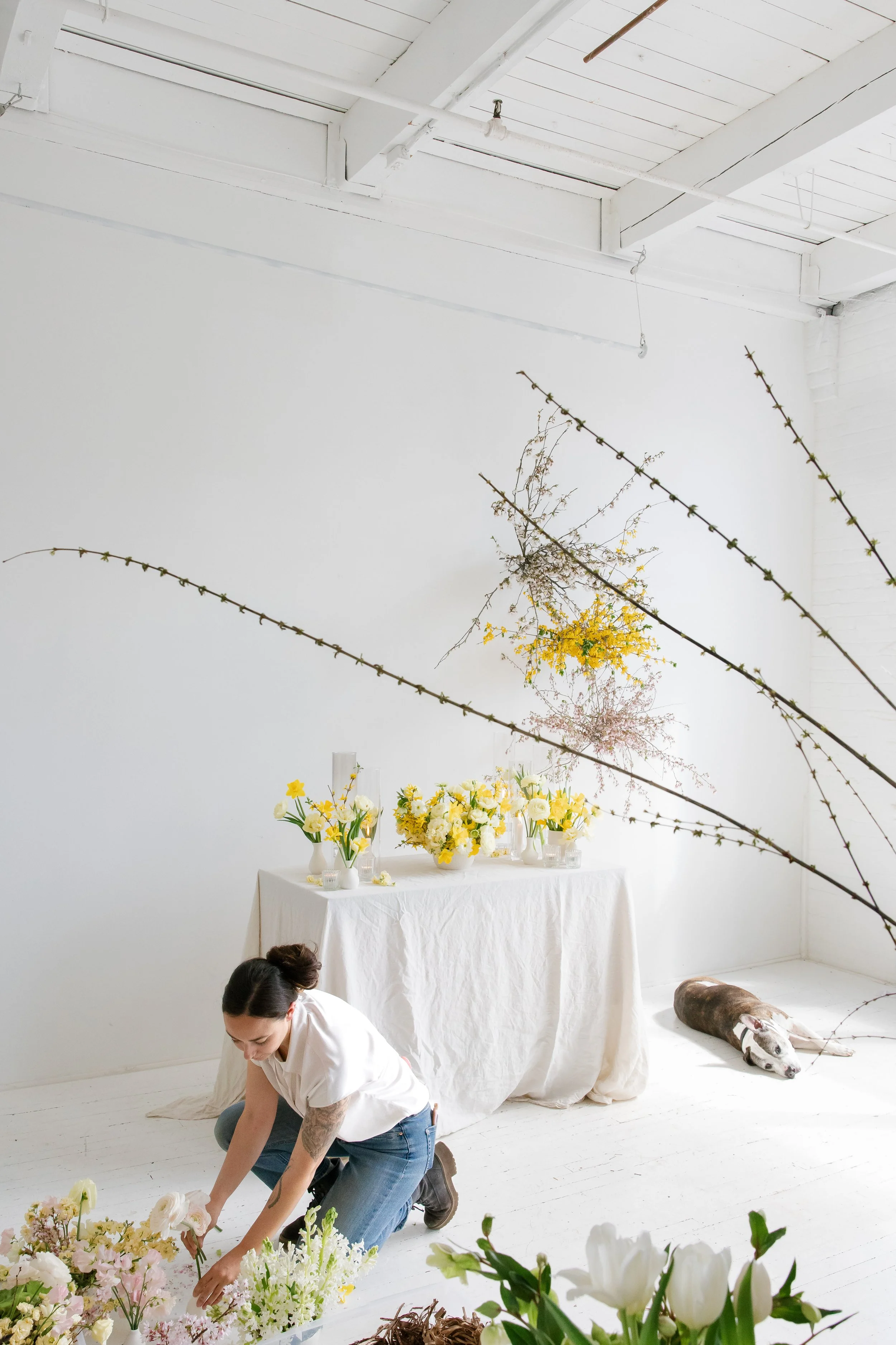 A woman arranging flowers on the floor in a bright white room with a table decorated with yellow and white floral arrangements and a white cloth cover. There are large floral arrangements on the table and dried branches hanging from the ceiling. A do