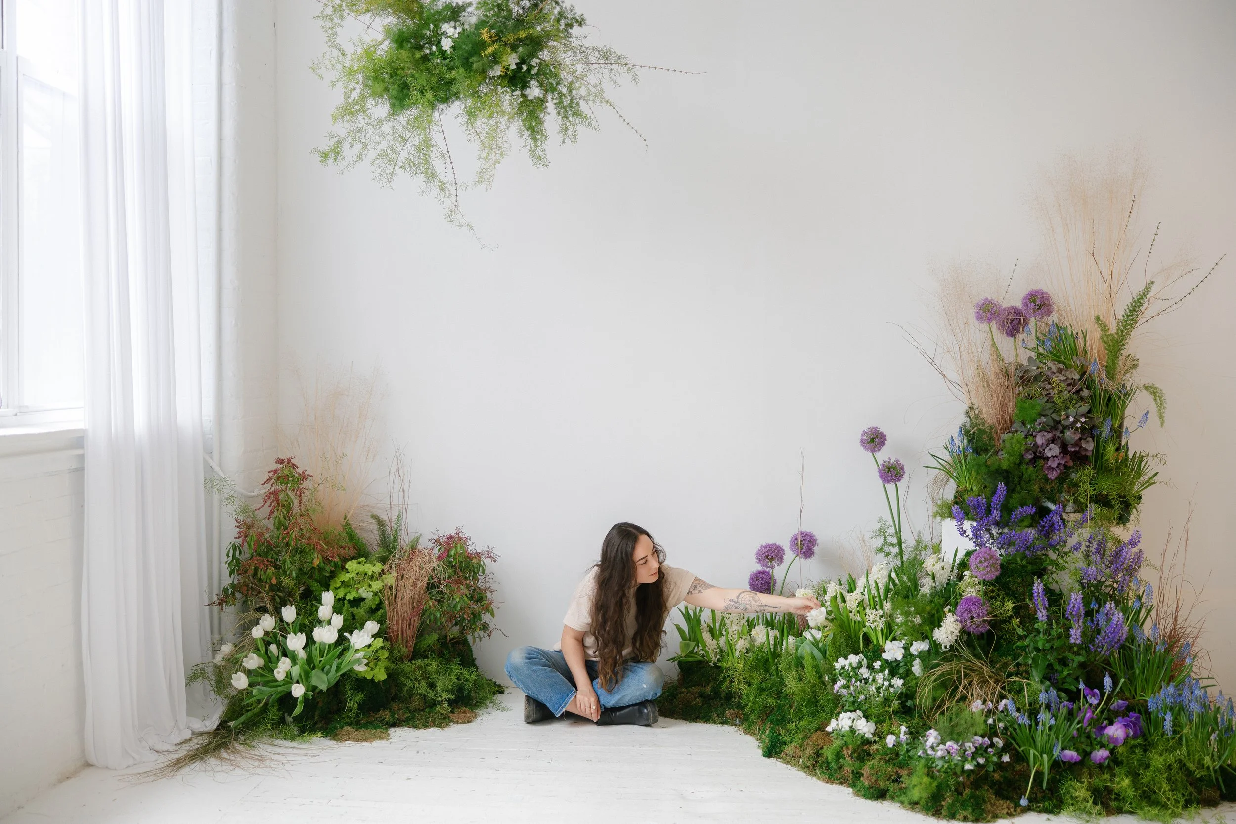 A woman with long dark hair sitting on the floor among various green plants and purple flowers inside a white-walled photographer's studio with a window and white curtains in The Alexander in Upstate, NY.