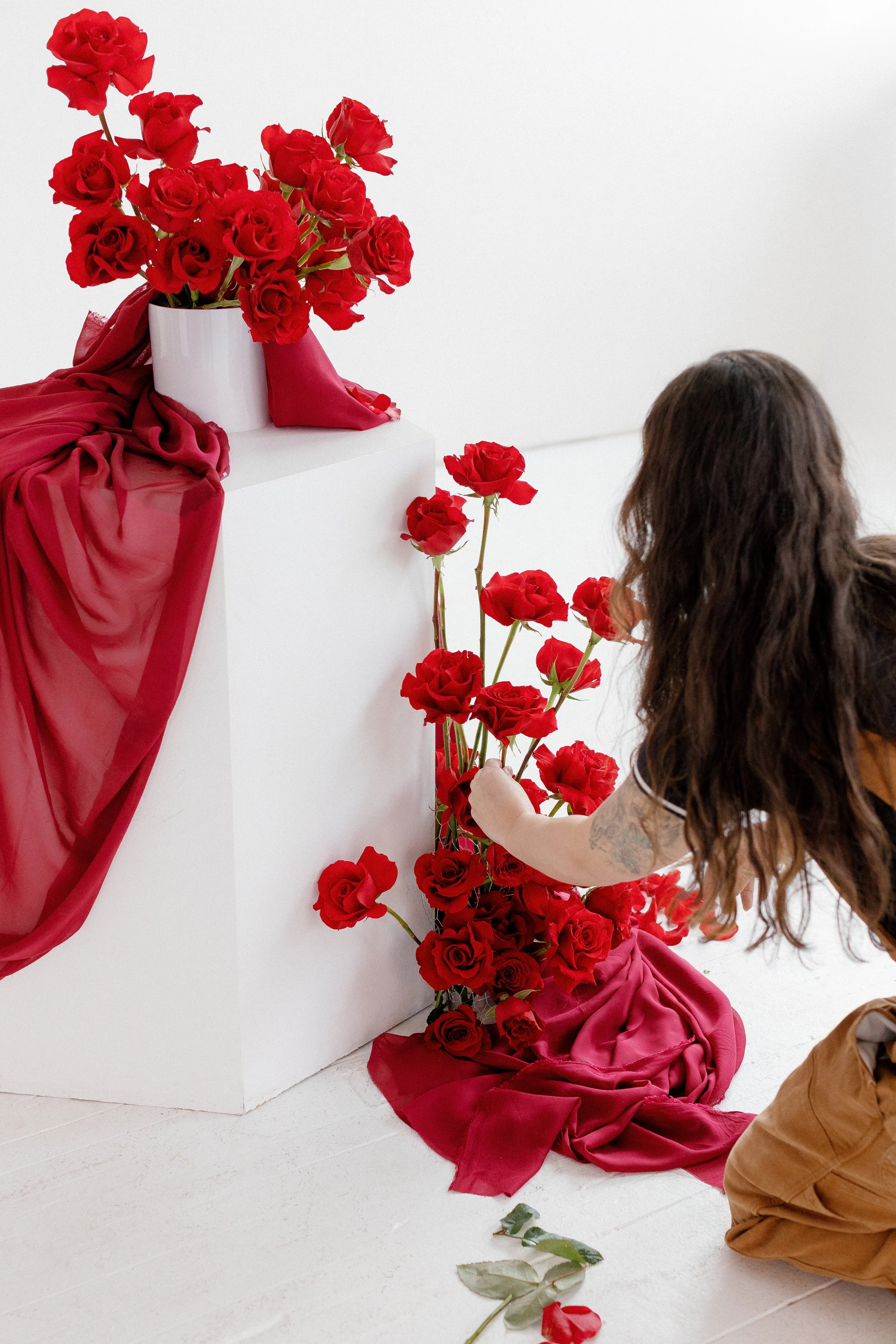 A person arranging red roses with a white vase on a white pedestal, decorated with red fabric, against a white background.