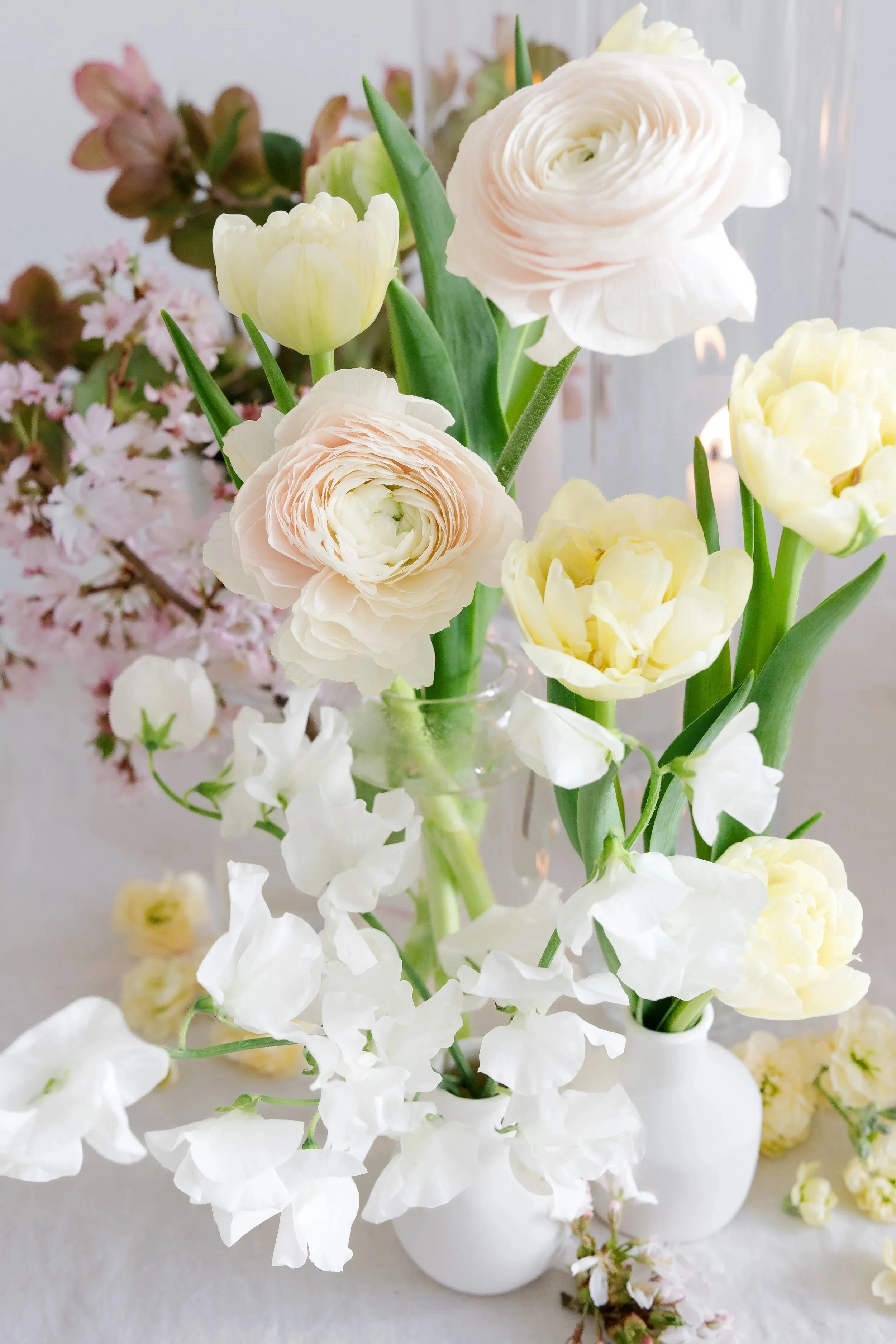 A close-up of a floral arrangement in white vases, featuring white and pale pink ranunculus, yellow and white tulips, and white stock flowers on a white surface with a soft background.