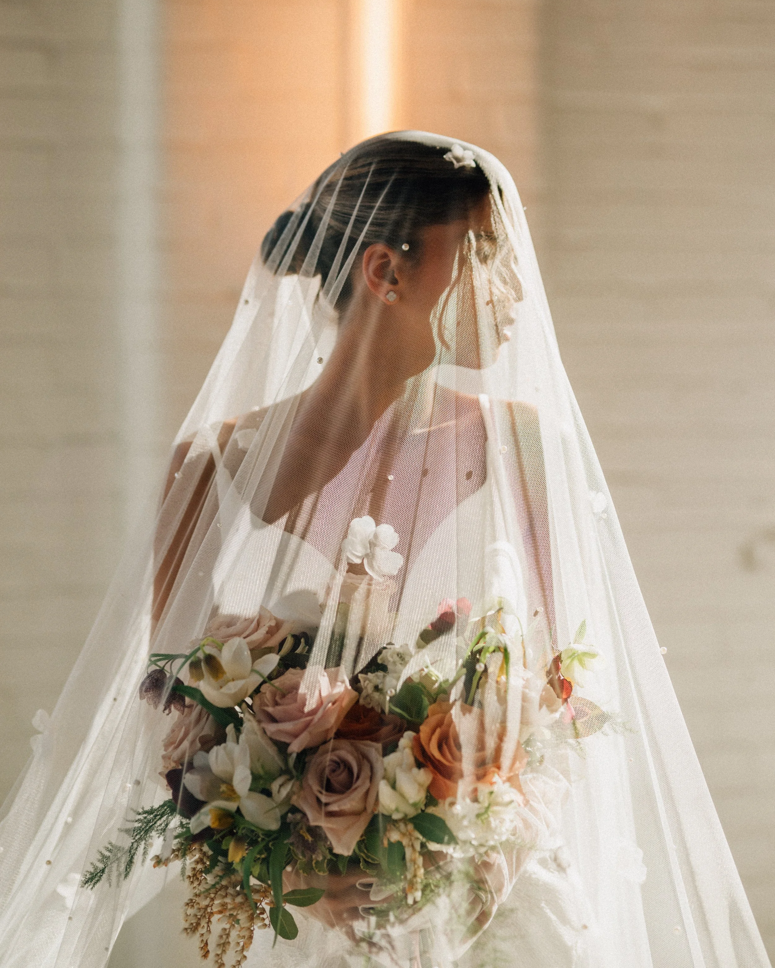 A bride with a veil and a high fashion bridal gown holding a moody bouquet off flowers including roses, hyacinth, hellebores, delphinium and more at Arbor Loft.
