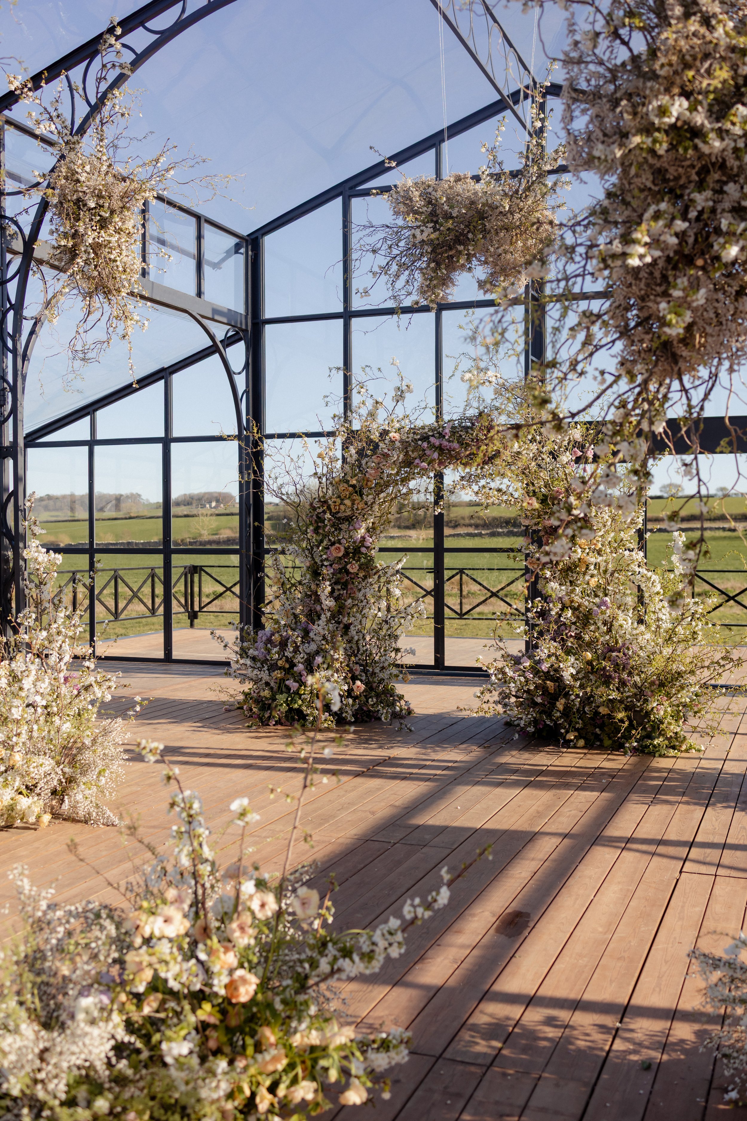 A greenhouse with glass panels surrounded by blooming white and pink flowers, set in a rural landscape with green fields and a clear blue sky at The Orangery with Trille Floral in Bath, United Kingdom.