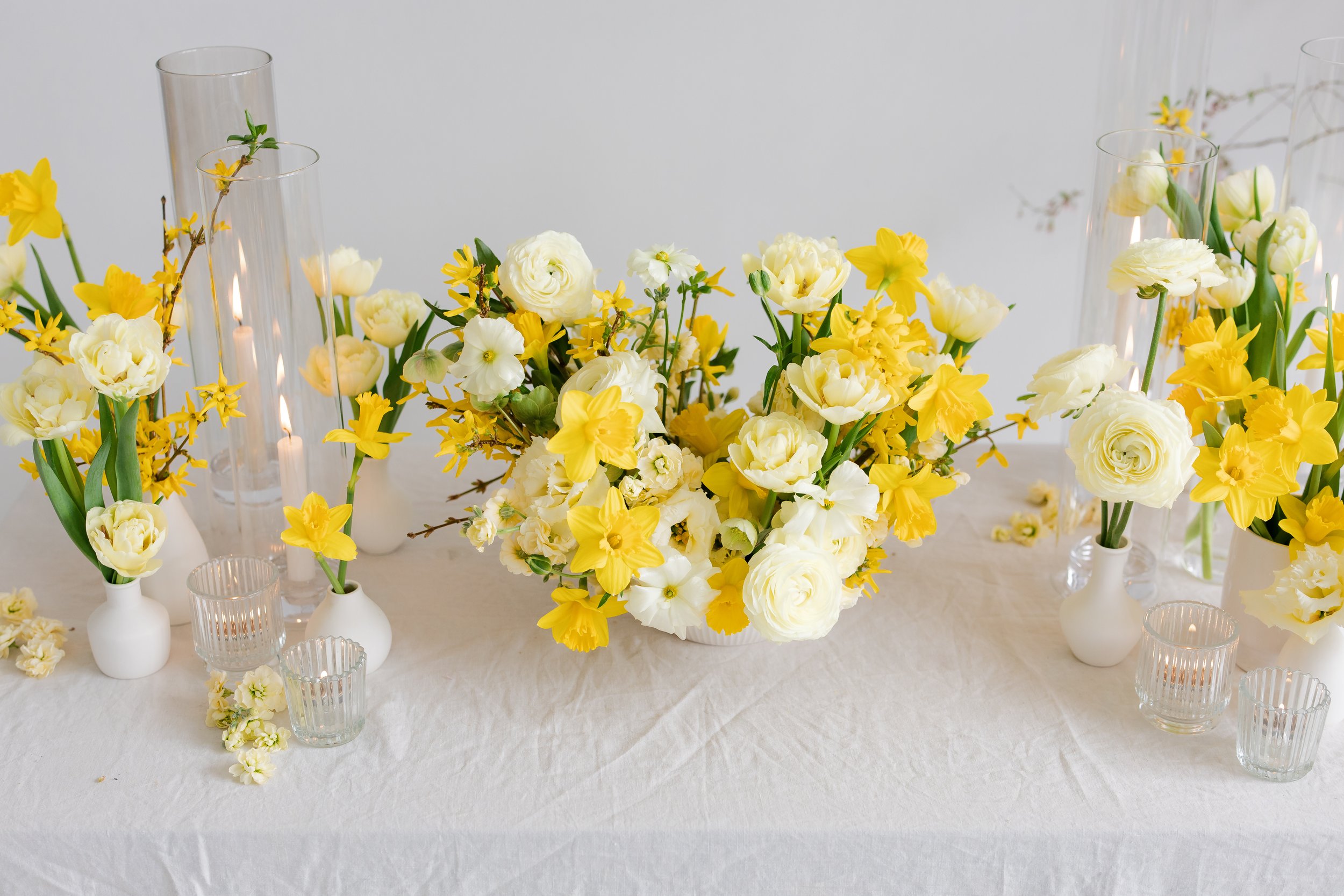 Table decorated with yellow and white flowers in white vases with glass candle holders around, set on white tablecloth.